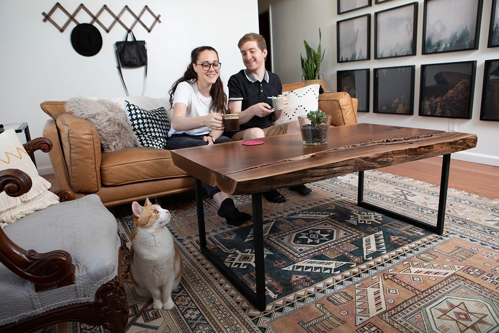 A couple sit on a warm brown leather sofa in front of a wooden coffee table. A cat sits in the foreground looking towards a wall of framed photos