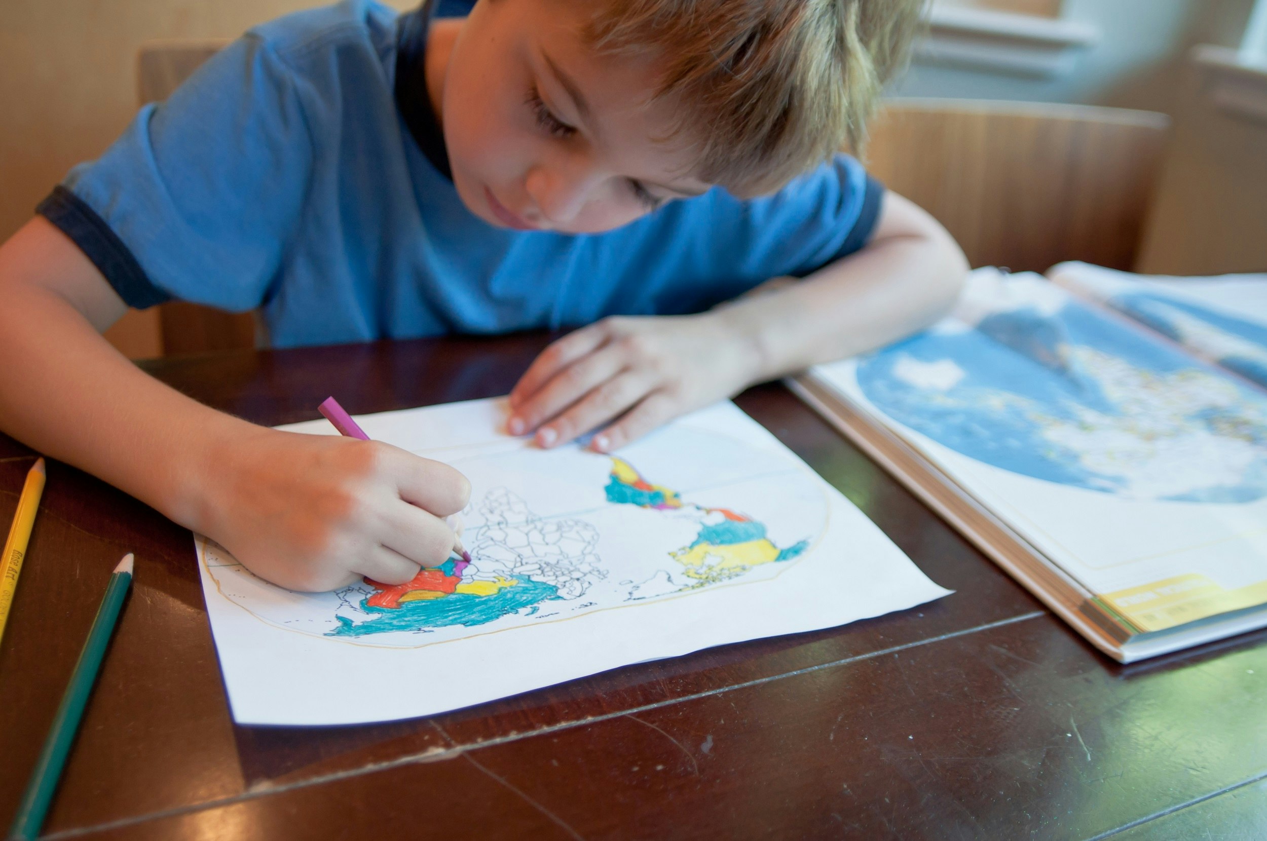 A young boy leans over his world map that he's colouring in. Next to him is an open atlas