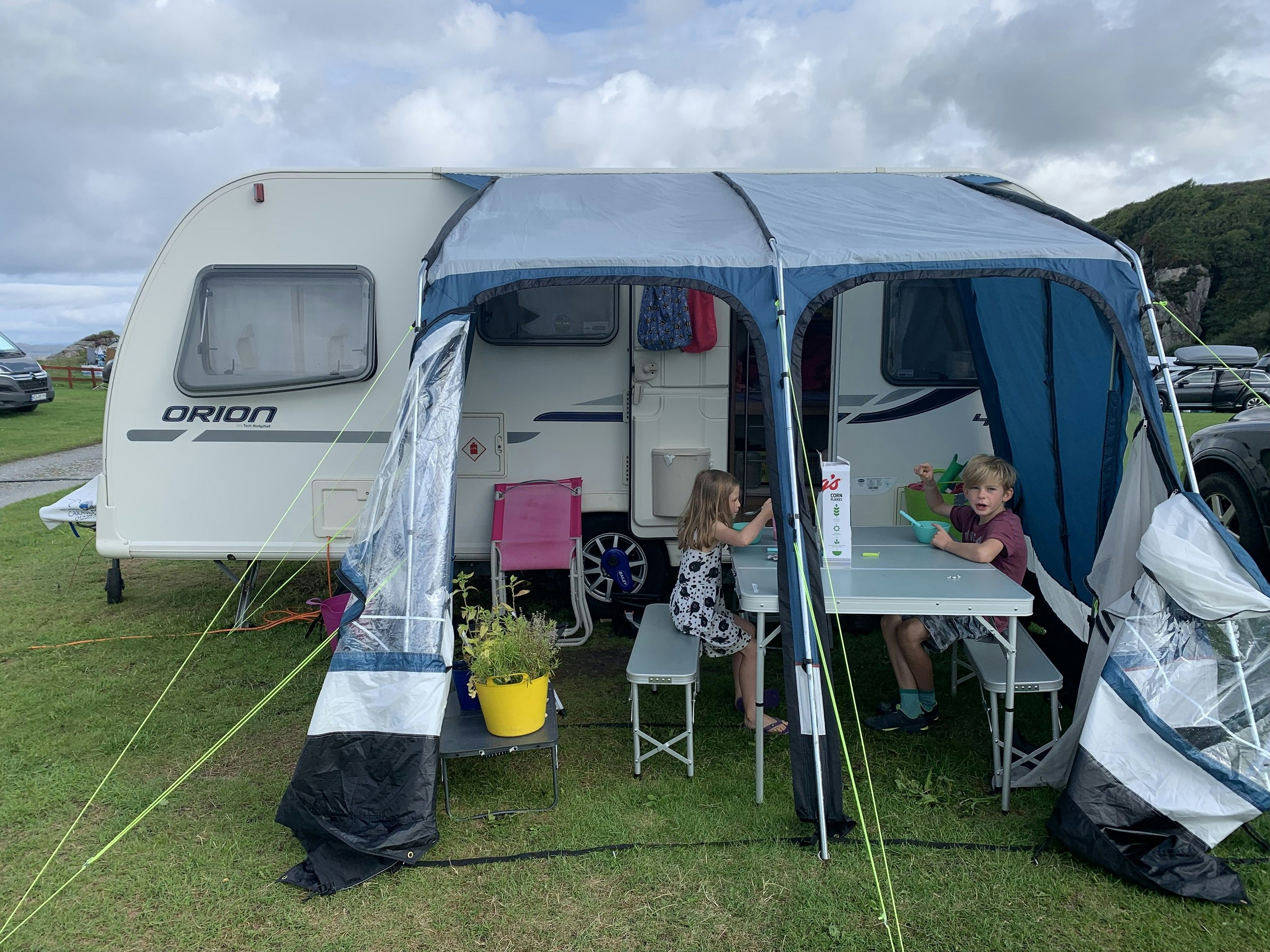 A caravan pitched at a campsite; an awning comes out from the side of the caravan; two children sit at a table under the awning eating breakfast.