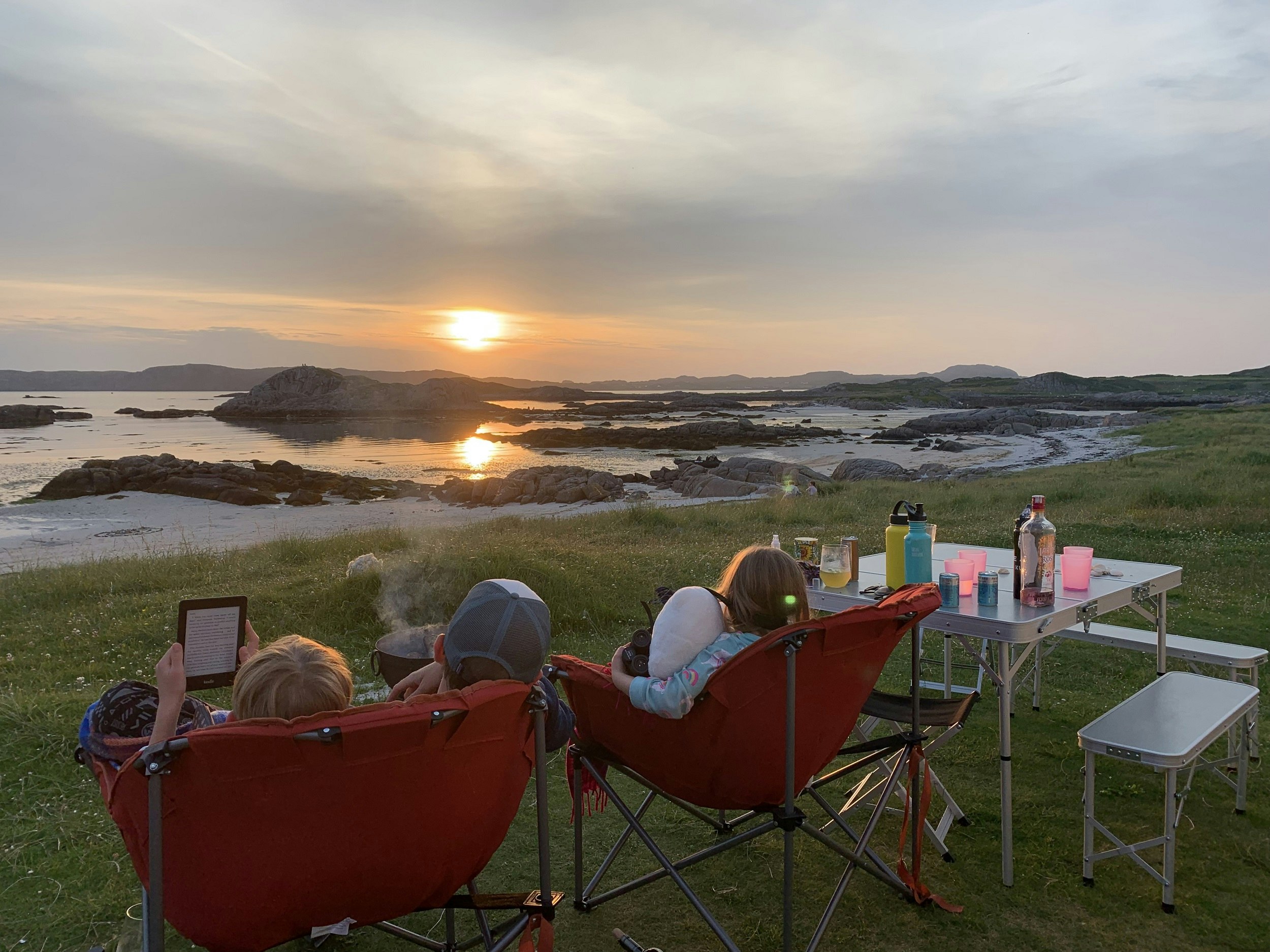 A dad and two children sitting in camping chairs watch the sun set over the sea.