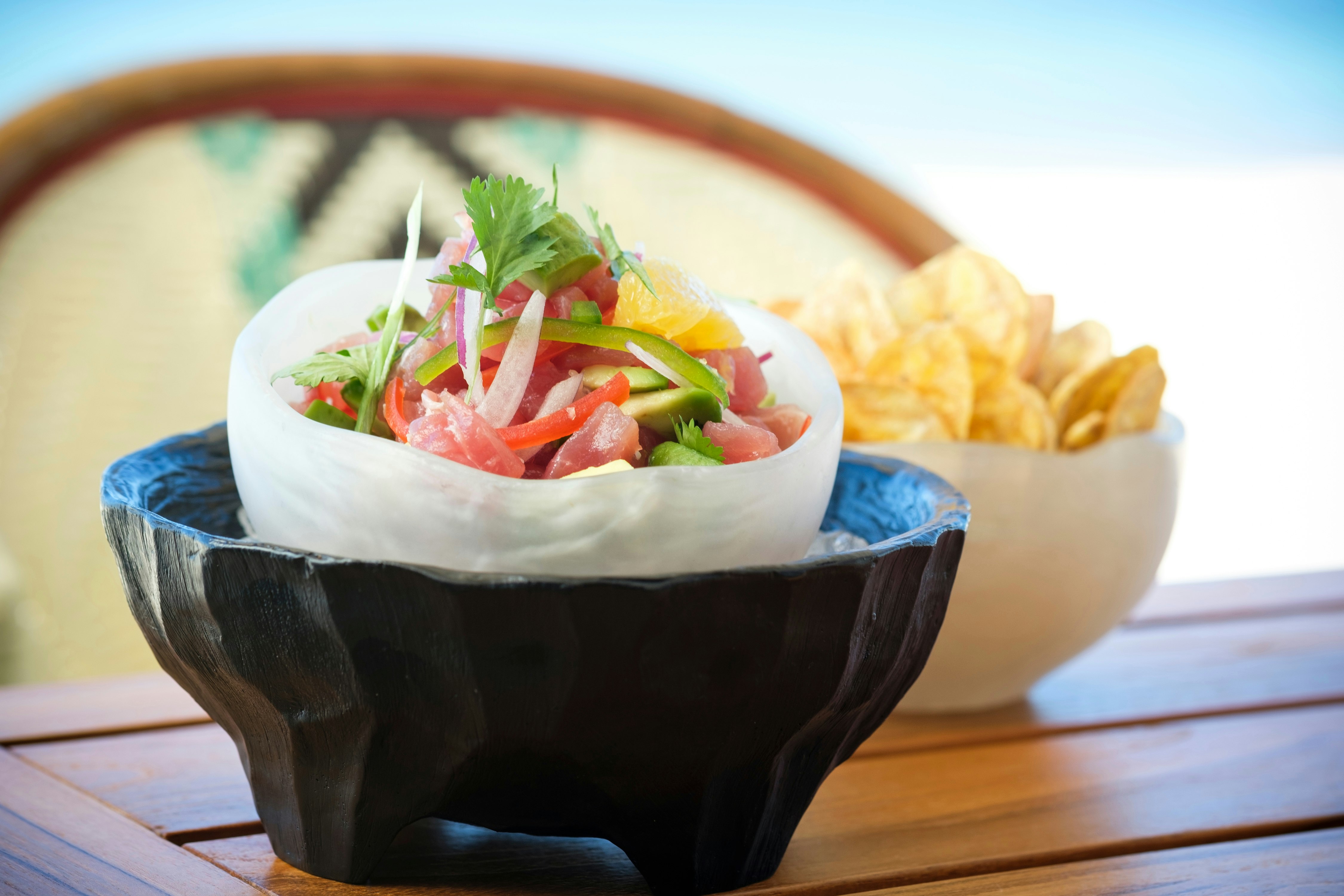 Two bowls on an outdoor table; in the background is a bowl of potato chips, and in the foreground is a bowl filled with a colorful, artistically presented salad.