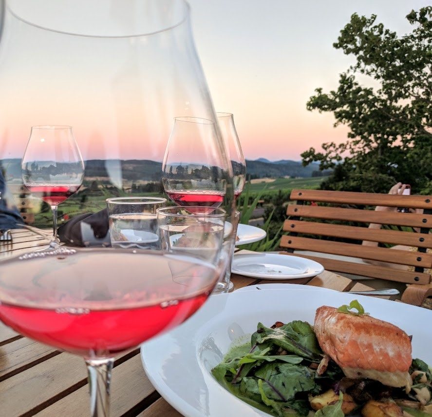 Three glasses of bright red wine in large tulip-shaped glasses stand out against a pink and orange sunset over the mountains in Oregon. In the foreground by one of the glasses is a white dish with a piece of salmon on a bed of greens. Further back on the table are a few empty dishes and a wooden bench