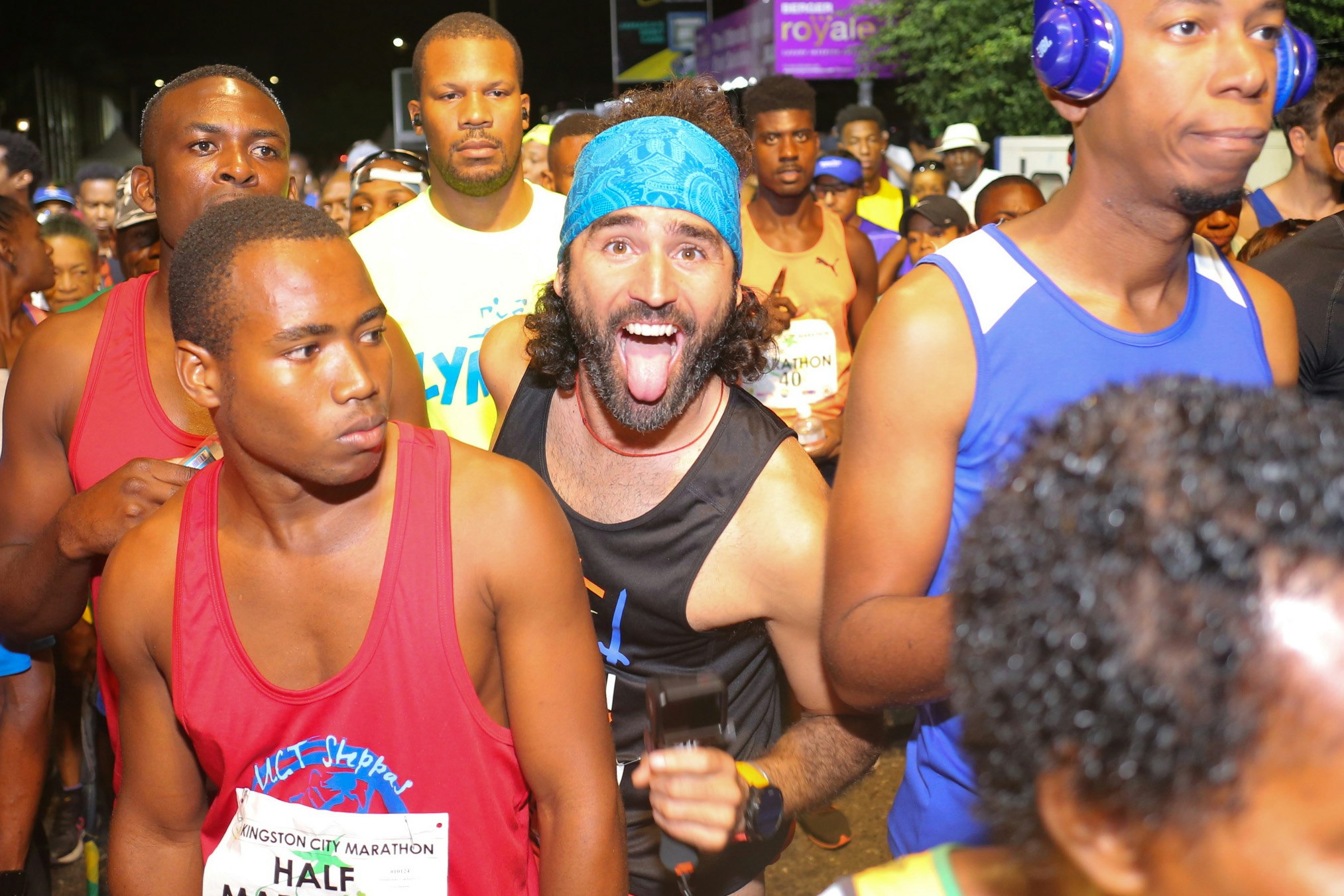 A crowd of runners standing in the flood lights at the start line of the marathon; one runner beams with excitement and his tongue hangs out from his wide-open smile