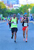 Two runners, one competing in the half-marathon, the other the full marathon, head down a flower-lined street; another runner who has finished cheers them on from the side of the street.