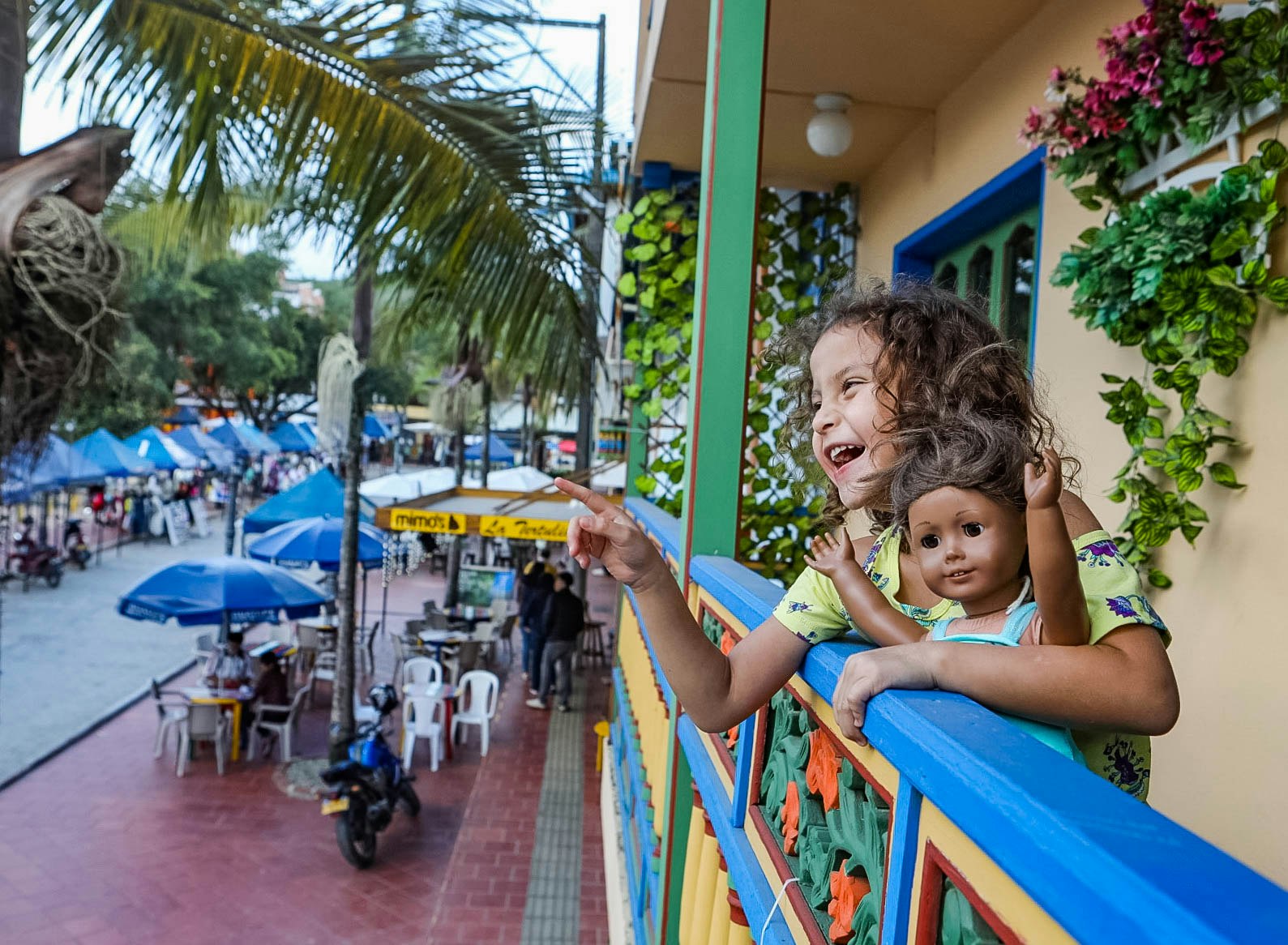 A little girl with curly brown hair and an American Girl doll who has the same light brown skin tone and wavy brown hair laughs and points to something just out of the frame. She is on a blacony with blue, green, teal, and orange railings, with tan stucco walls behind her and a shelf overflowing with vines and flowers. On the street below are long rows of blue tents over street vendors and blue umbrellas over white plastic tables and chairs for nearby cafes. Palm trees hang over the brown tiled sidewalks