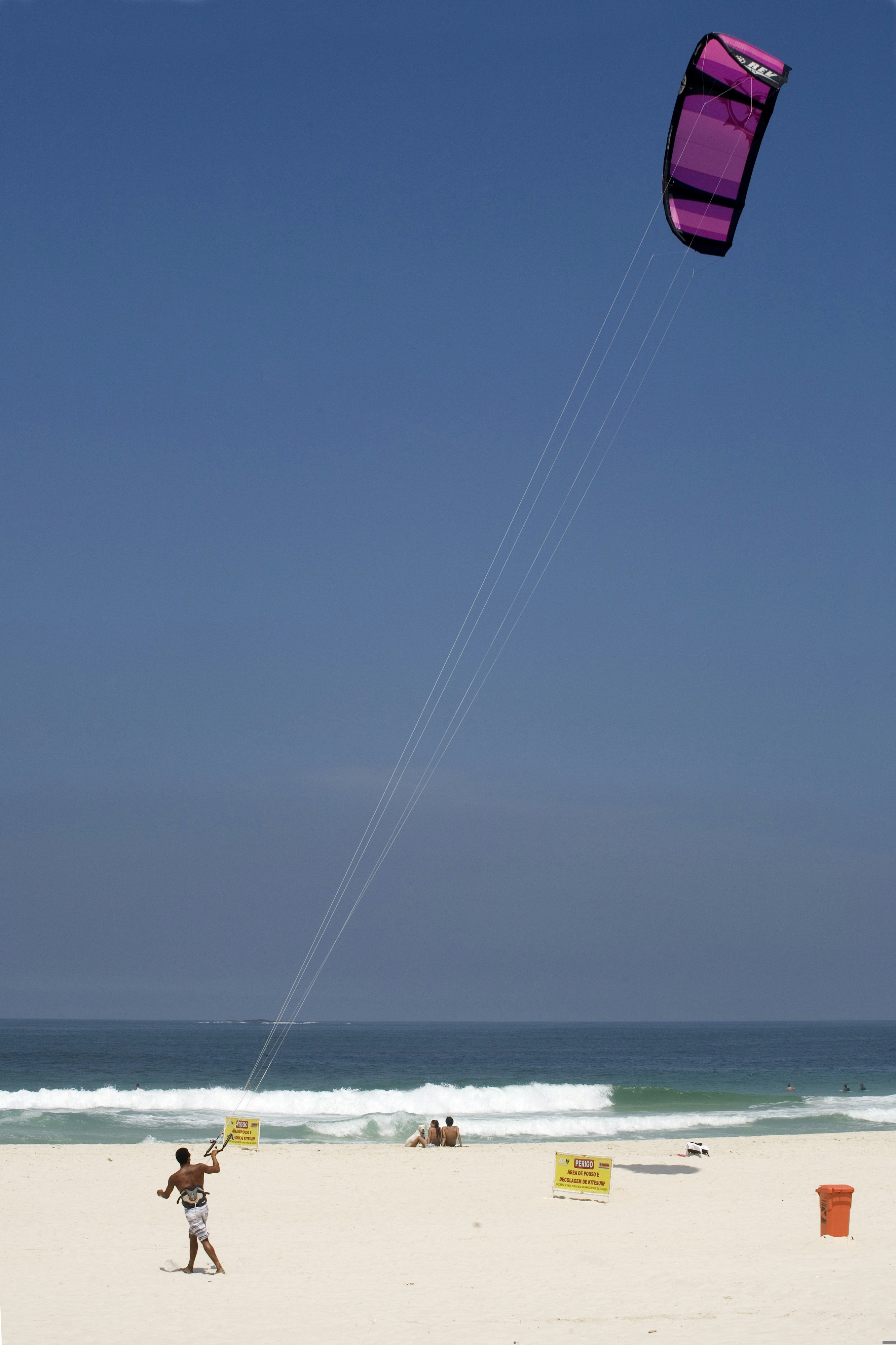 A kitesurfer walks along the sand at Barra da Tijuca beac with his purple kite flying high above; a couple of other beach-goers sit in the sand.