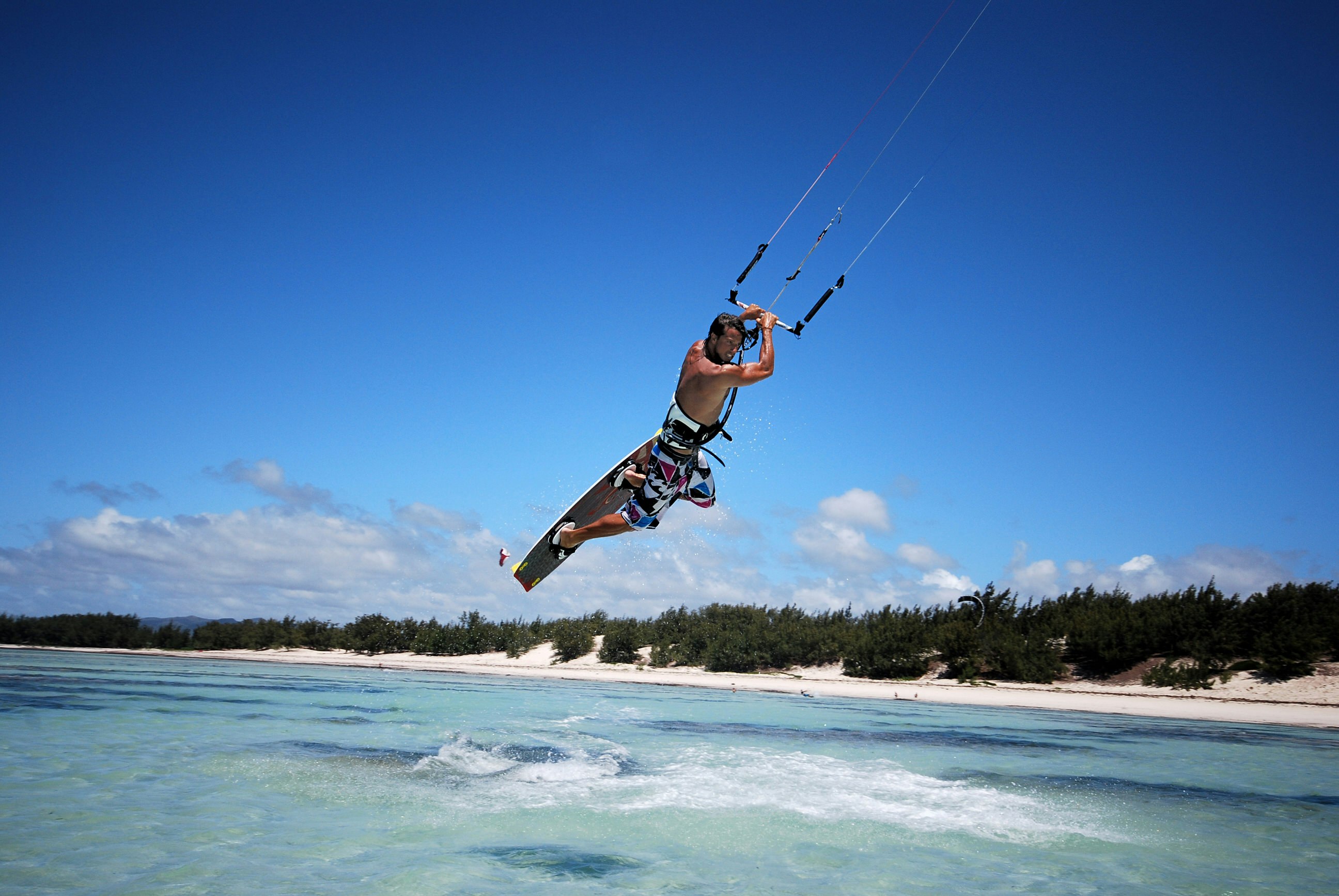 A kitesurfer sails through the air, his kite above him and out of the picture; below him is tropical waters, with a distance beach and forest in the background.