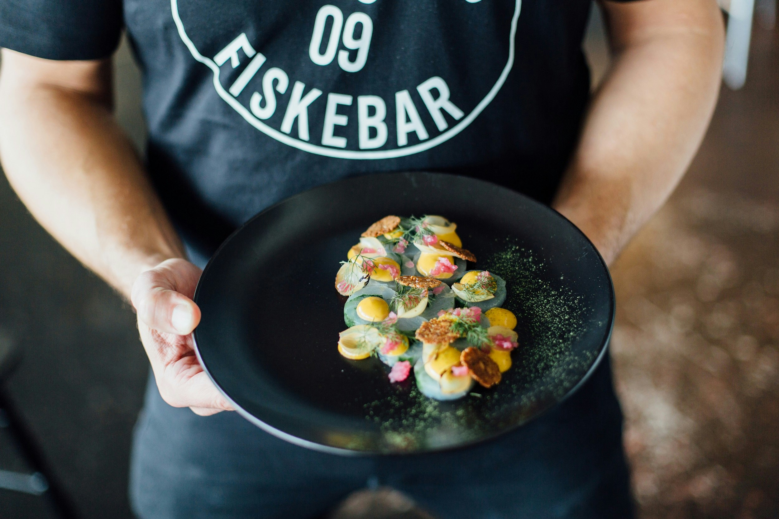 A man wearing a black T-shirt emblazoned with Fiskebar holds a black plate sprinkled with green shavings and colourful New Nordic food