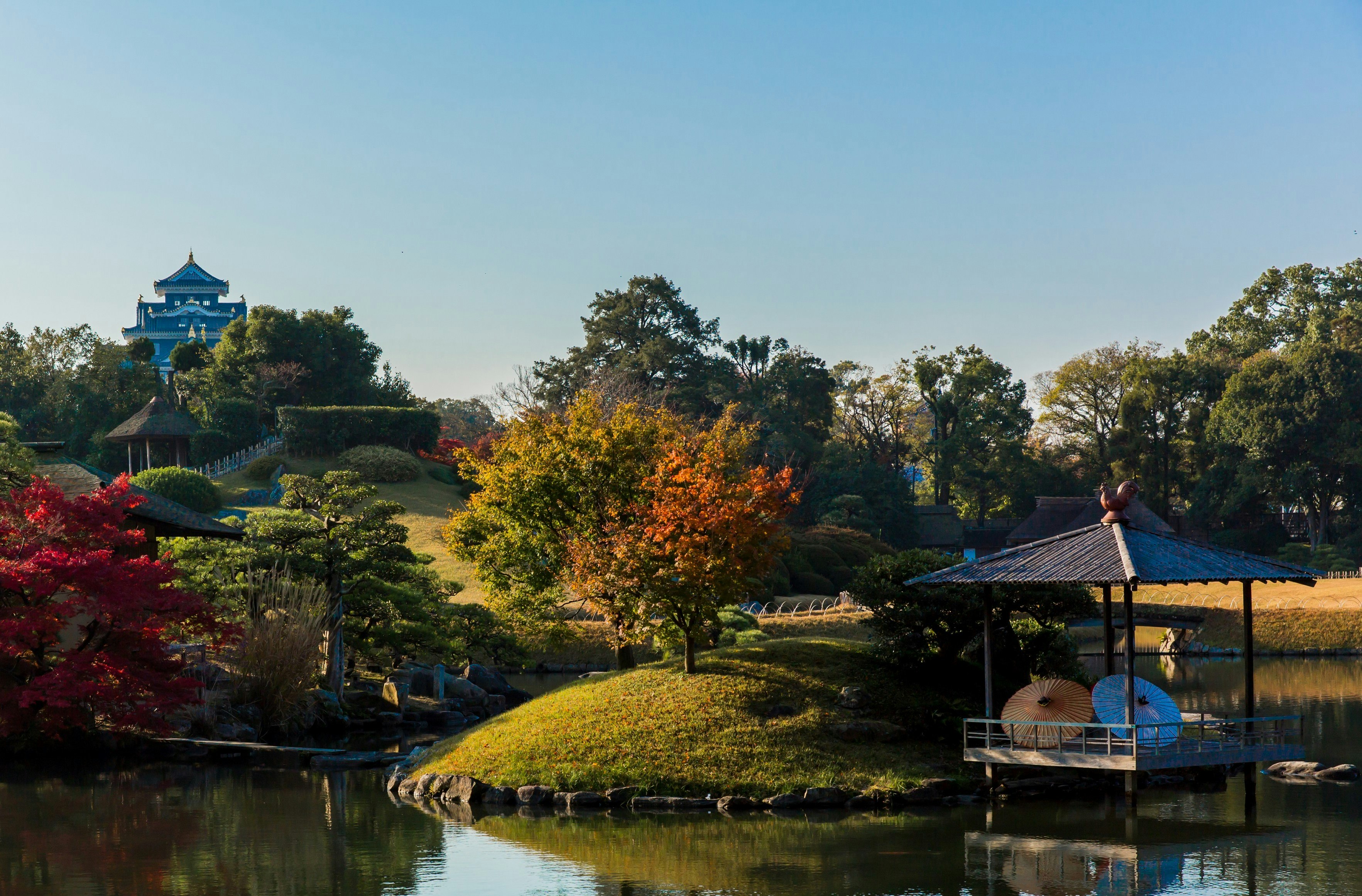 A view of Koraku-en garden in Okayama, with the still lake in the foreground leading away to green hills dotted with trees and an ornate castle visible in the background.