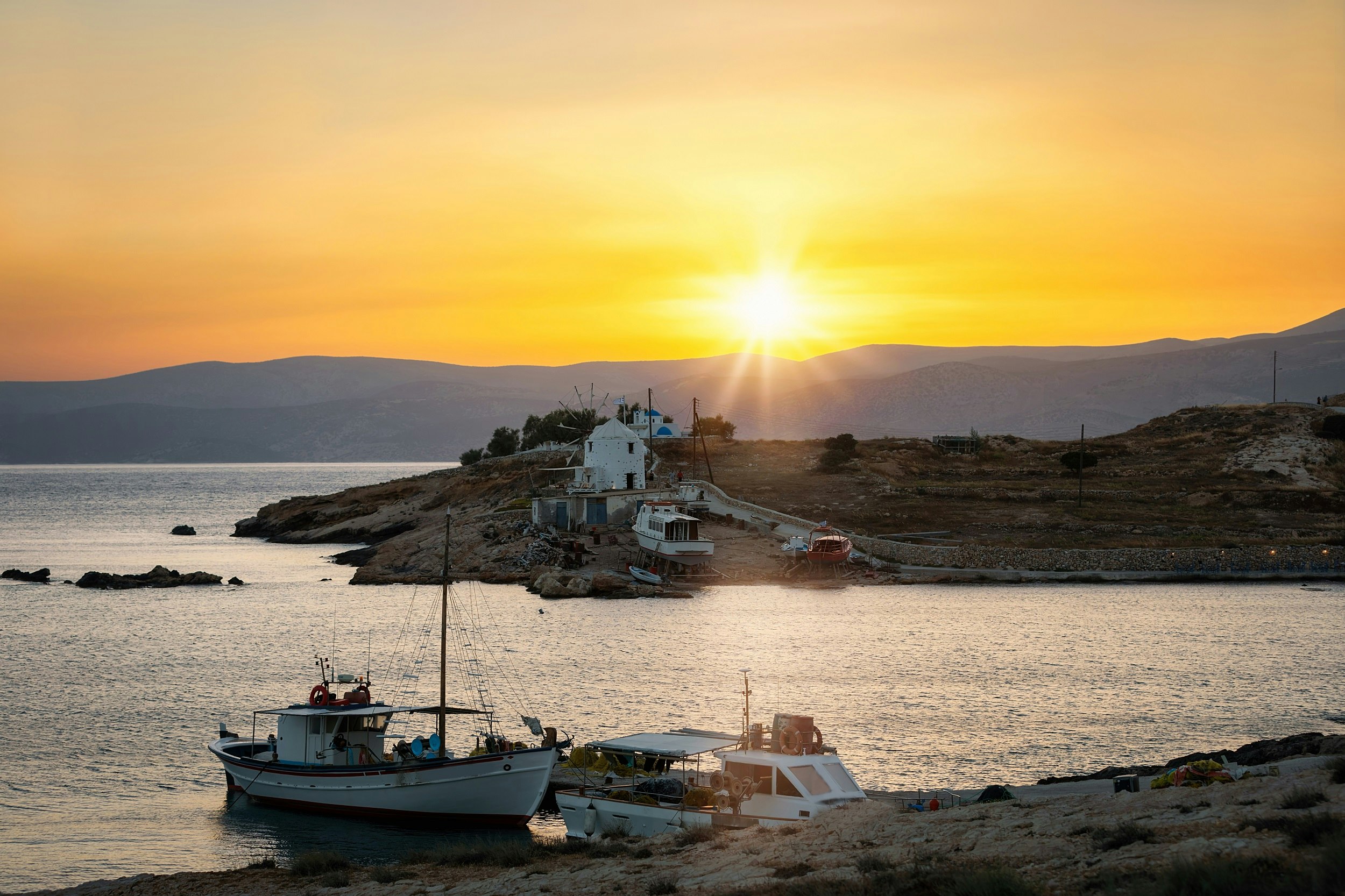 Two small fishing boats are docked near a rocky shore. A single white building stands on the shore. The sun sets over the hills in the background.