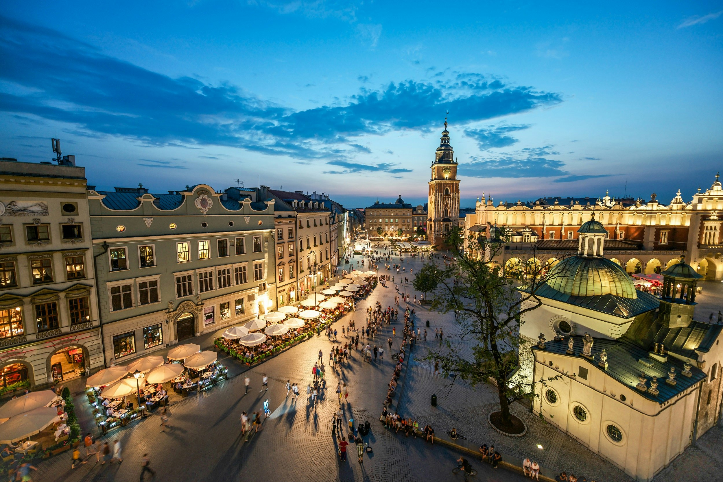 A view down over the square lined with grand buildings. The Town Hall Tower is the tallest of them all. Many outside tables at restaurants are occupied as the last light from the sun fades in the sky
