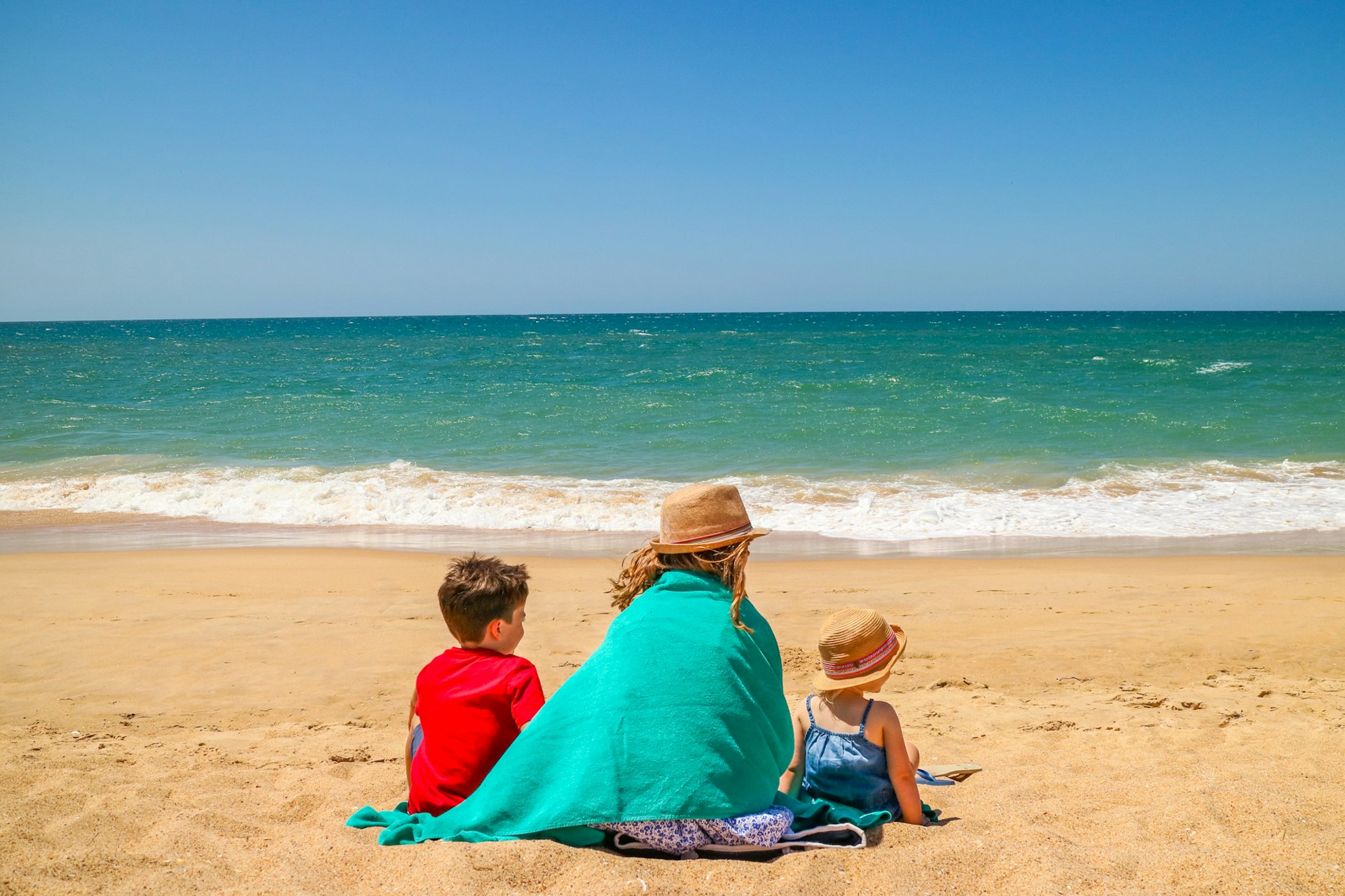 A woman sitting on a sandy beach between two children; they all have their backs to the camera and are looking out towards the sea.