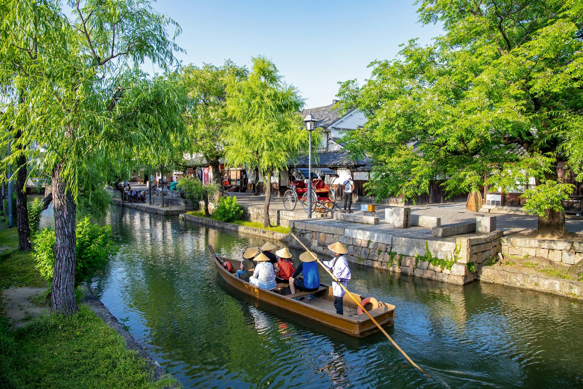 A man in a kimono rows a boat of passengers, all wearing conical rice hats, along a narrow river in the town of Kurashiki. The river is flanked by green trees.