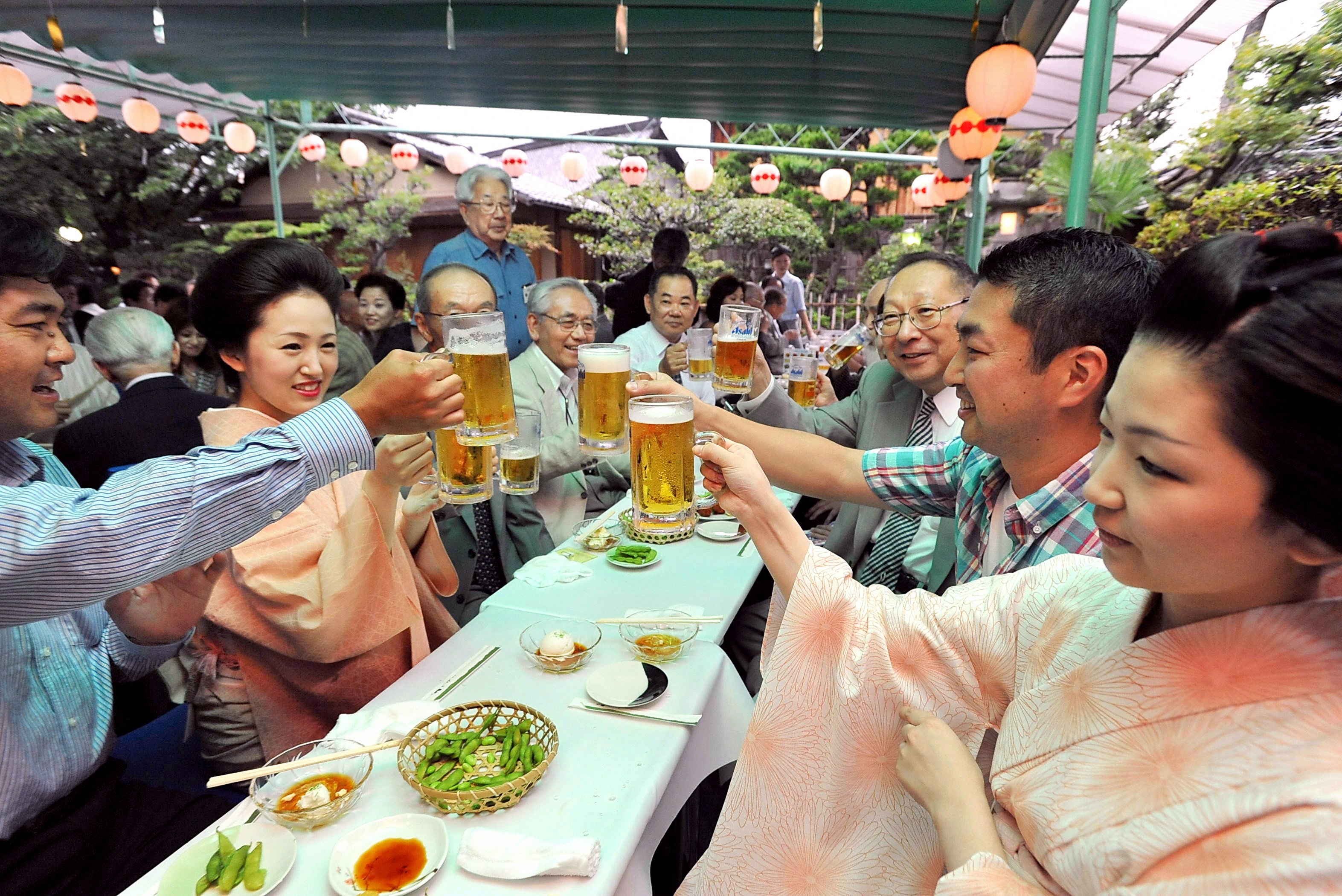 A narrow table with a white cloth and small dishes of food sits between two rows of people, some men in button-down shirts and some women in light pink kimono, who are all hoisting tall steins of beer in a toast at the Kamishichiken Kaburenjo Theatre in Kytoo