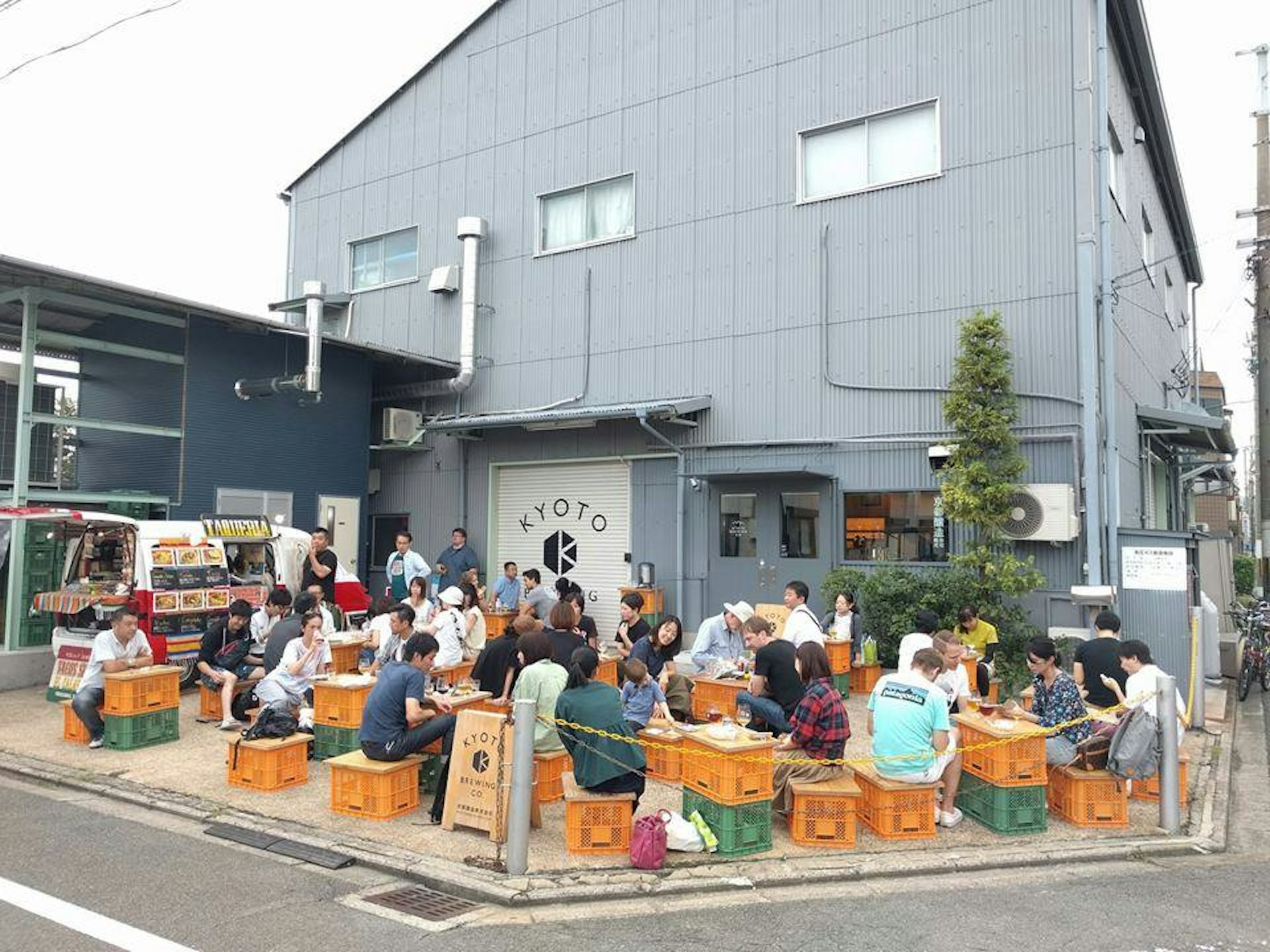 A patio with plastic crates for chairs and tables is full of patrons enjoying their beers and chatting with one another against the backdrop of a grey corrugated metal building