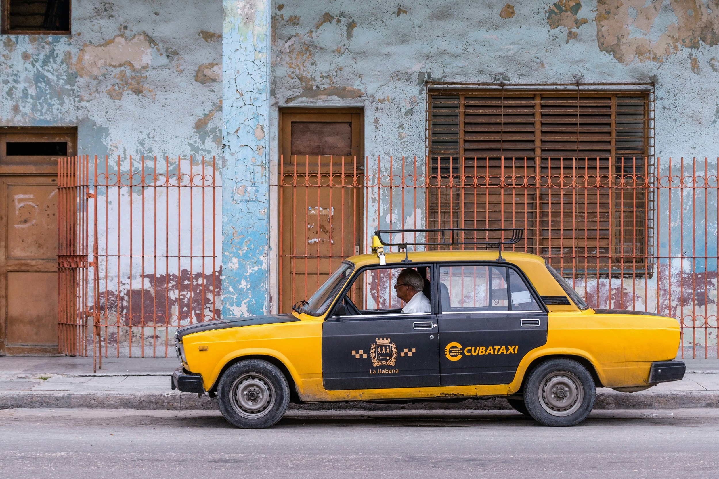 An old yellow Lada car, with its doors, hood and boot painted black, has CUBATAXI emblazoned on its rear door; it's driving past an older building with a barred fence surrounding it.