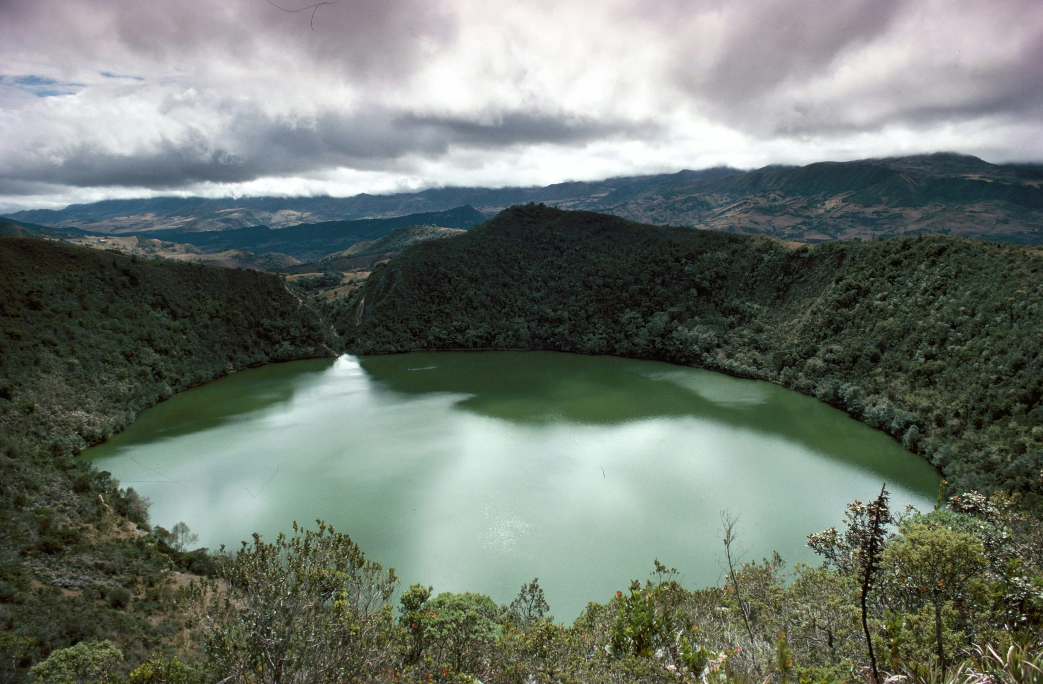 The brilliant green waters of Lake Guatavita contrast with the round cirque of mountainous terrain around the lake shot at a slight angle 