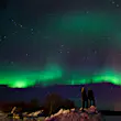 Two people watching the green streaks of the Northern lights across a dark sky