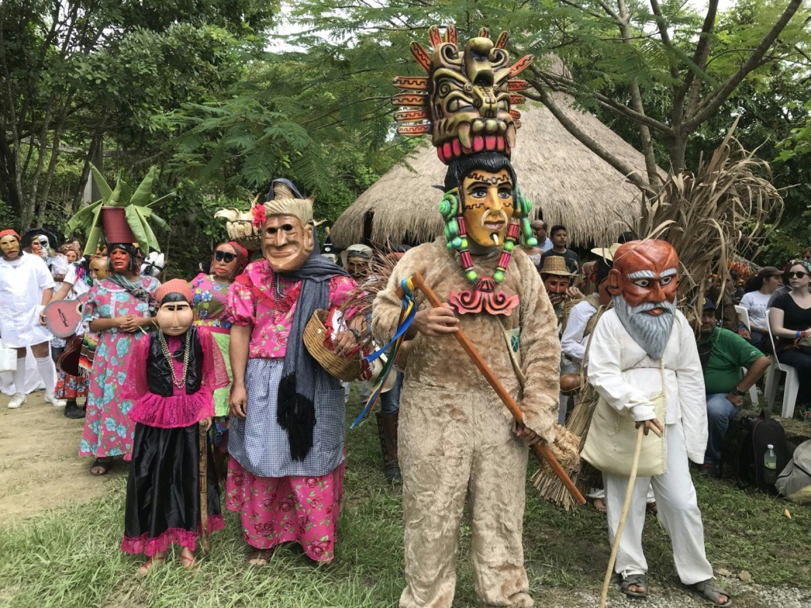 A procession of people dressed in traditional colorful garb and wearing large masks walk through a village; Xantolo