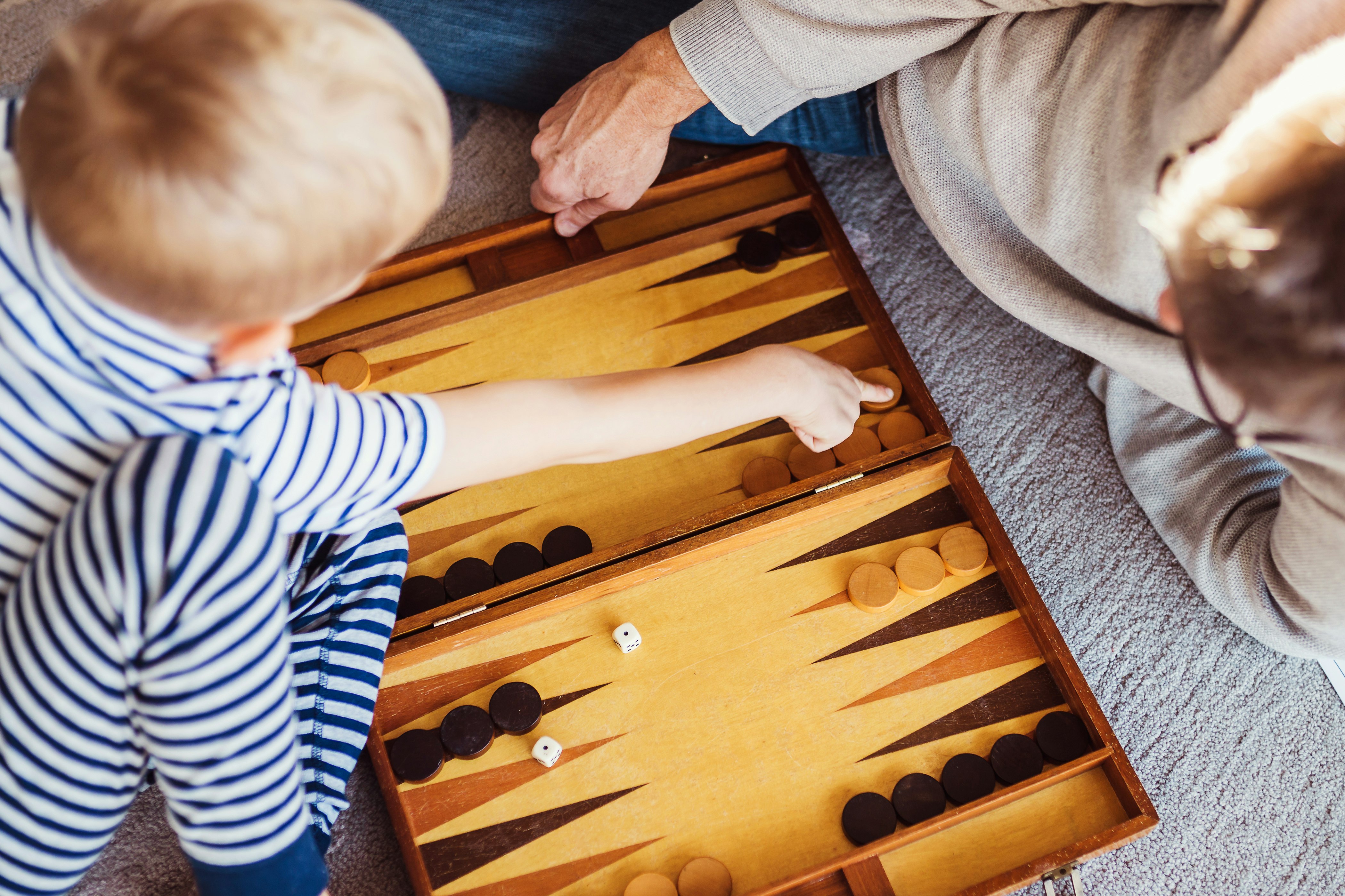 A young child is playing backgammon on the floor with their parent; the child is moving one of the pieces on the board.