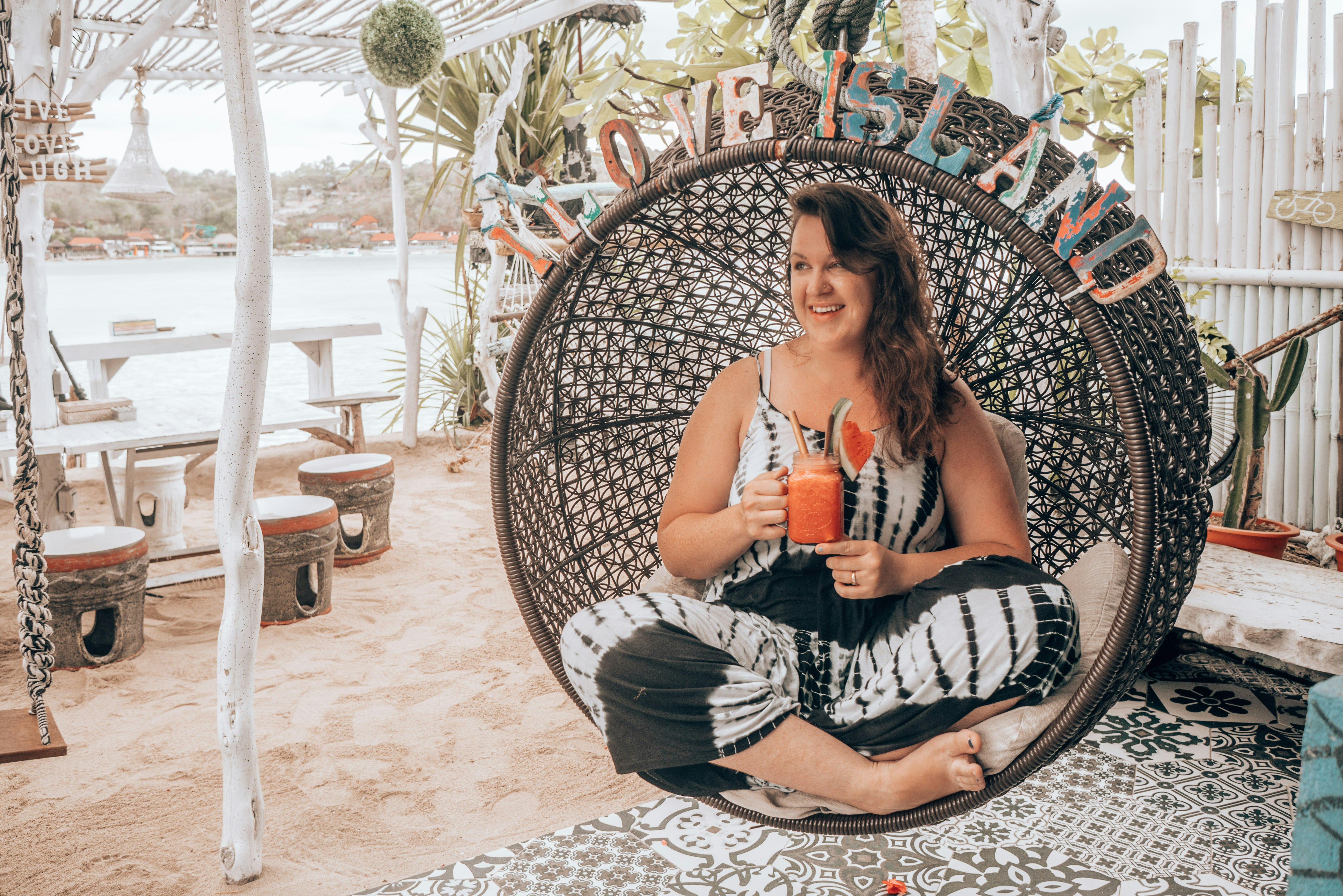 A woman with long medium brown curly hair sits in a wicker egg chair holding an orange juice in a mason jar. Letters in a distressed shabby chic aesthetic sit on the top of the chair reading I Love Island.