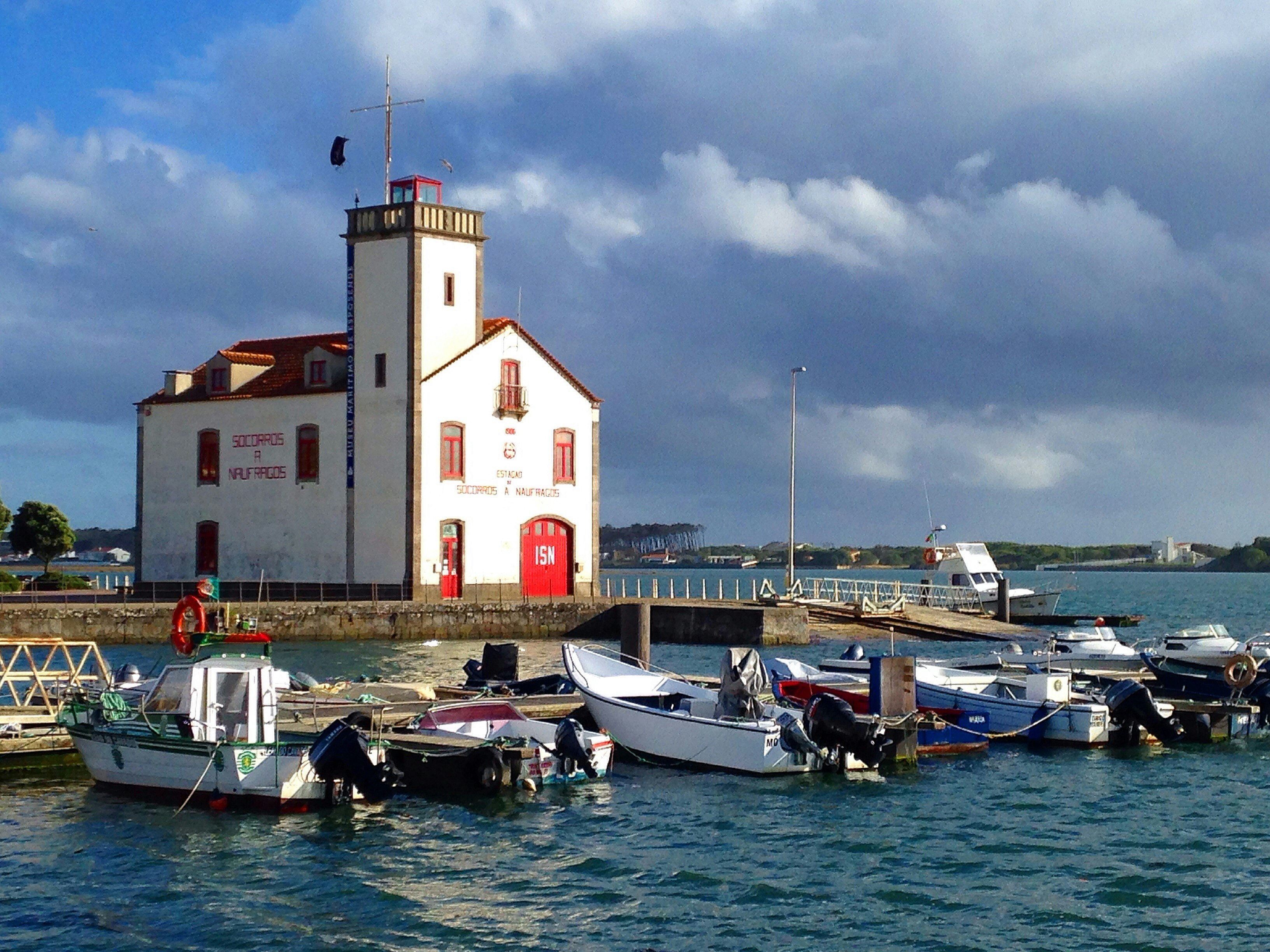 A simple white building with red-painted doors and windows stands at Esposende harbour on the Camino Portugués de la Costa route. There are several small boats bobbing on the water.