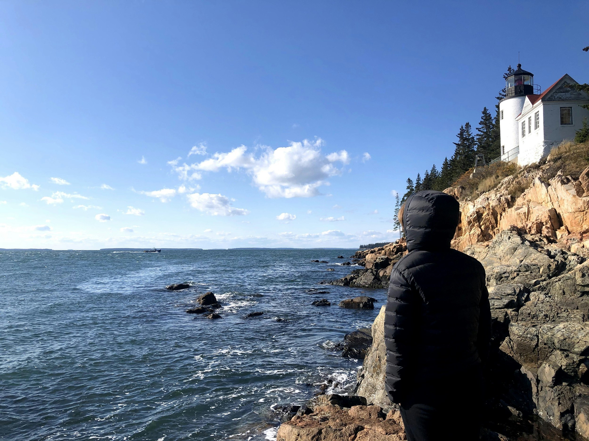 Lindsey Bathke stands in a black puffer coat on the rocky Pacific Northwest coast with a white lighthouse in the background  