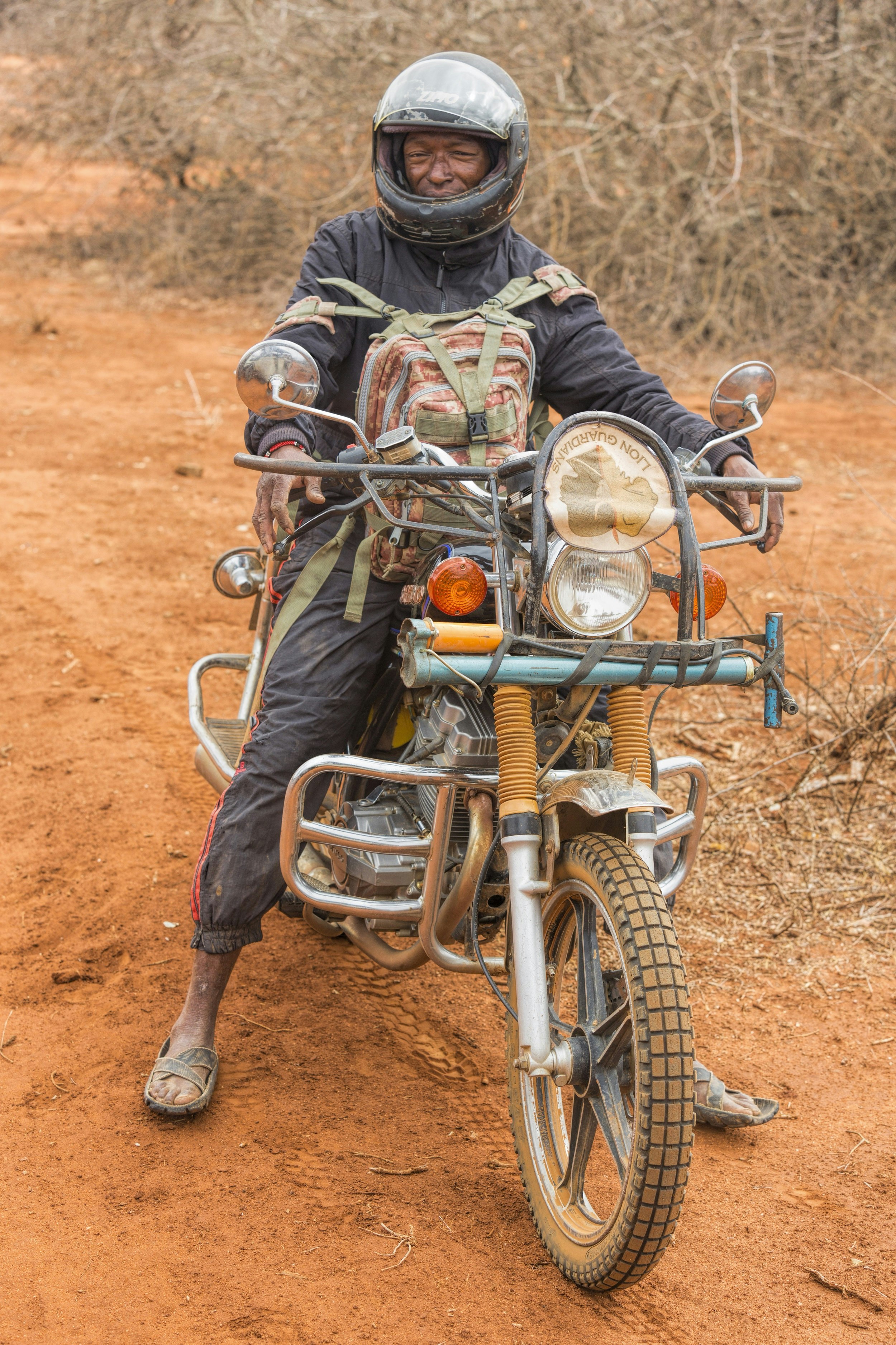 A Maasai warrior, clad in a black set of overalls, black helmet and sandals made of old tyres, sits astride a dirt-covered motorbike; he's wearing a backback on his chest, with the bike sitting stationary on a dirt road.