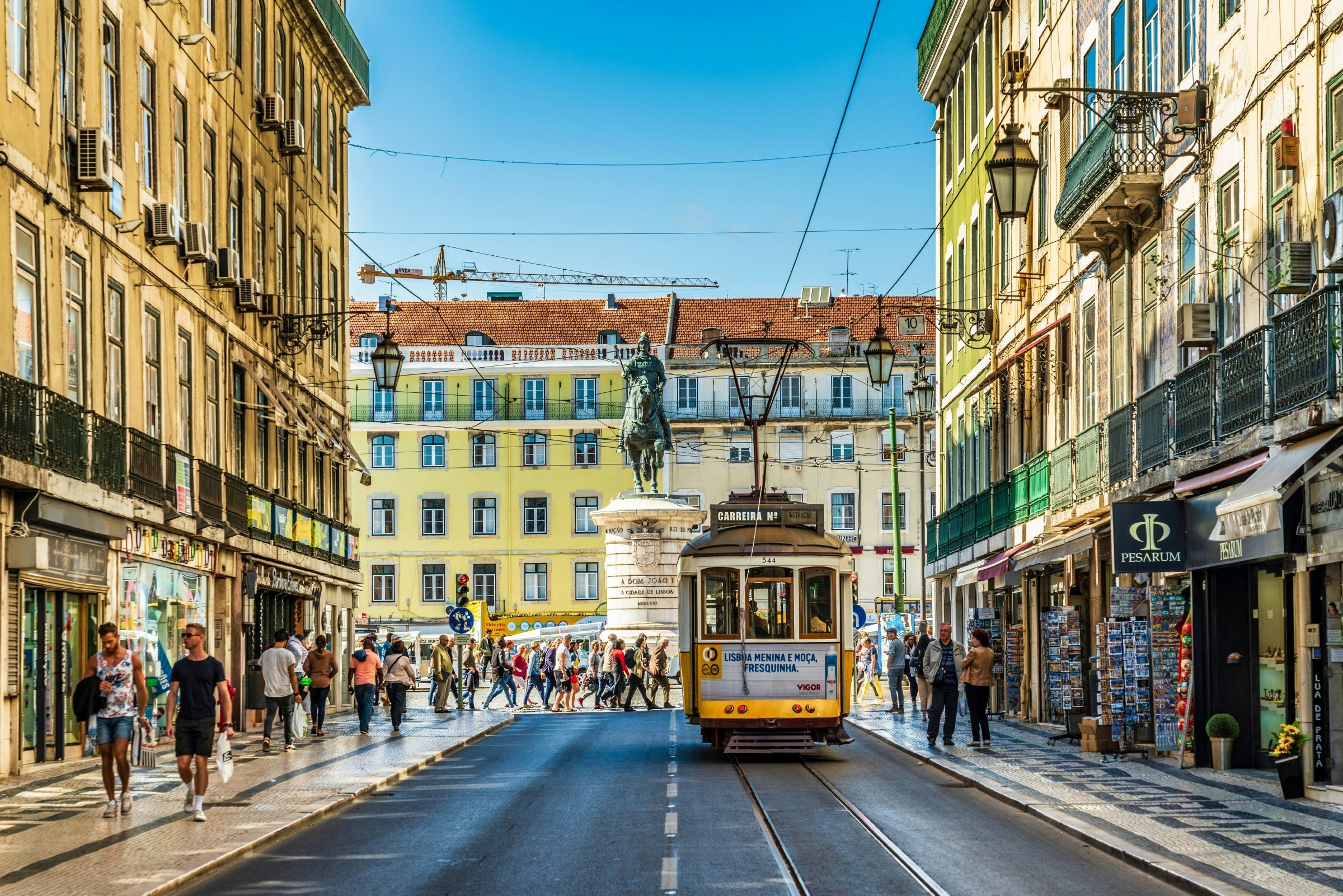A yellow tram is travelling down a busy shopping street in Lisbon, Portugal, on a sunny day.