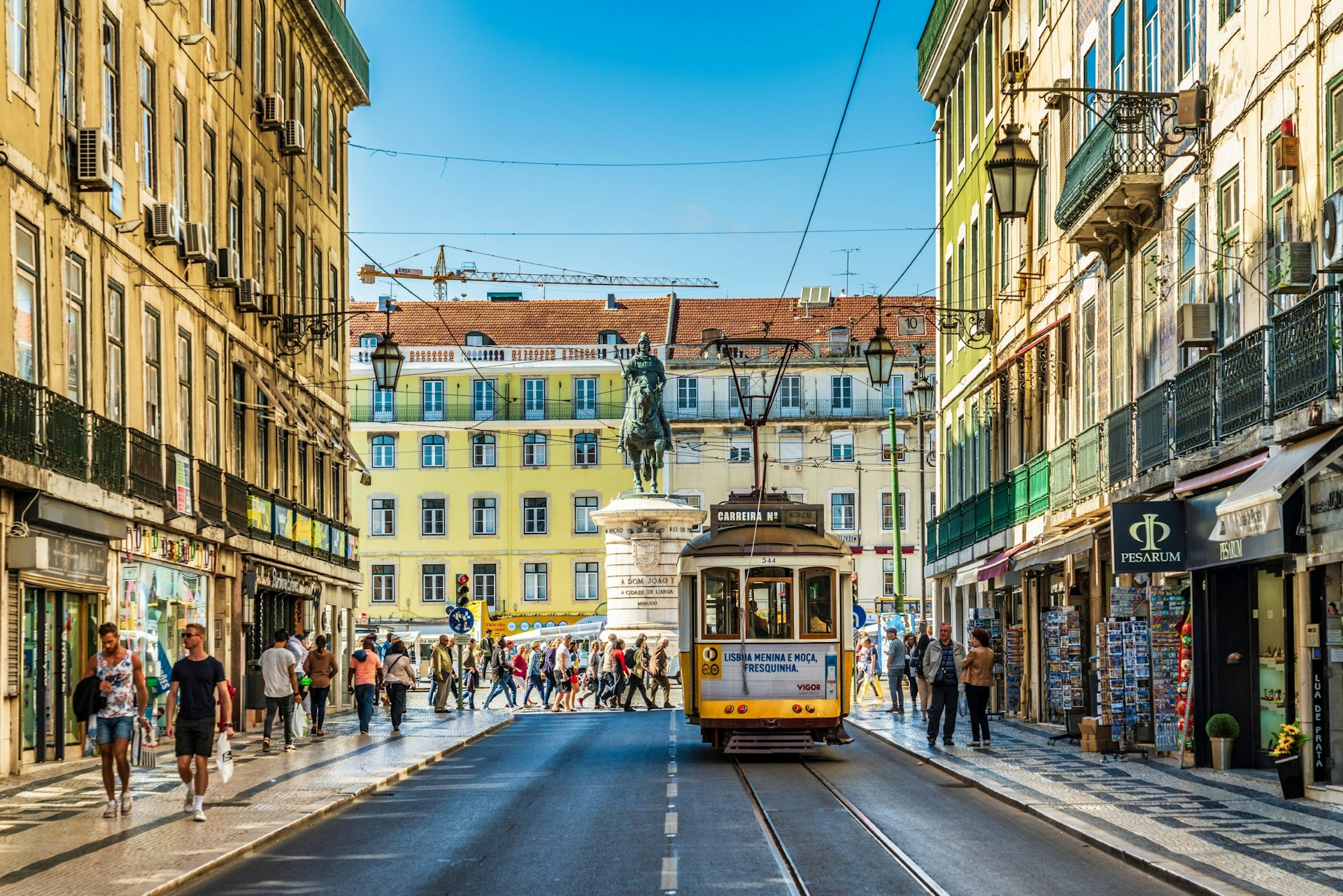 A yellow tram is travelling down a busy shopping street in Lisbon, Portugal, on a sunny day.