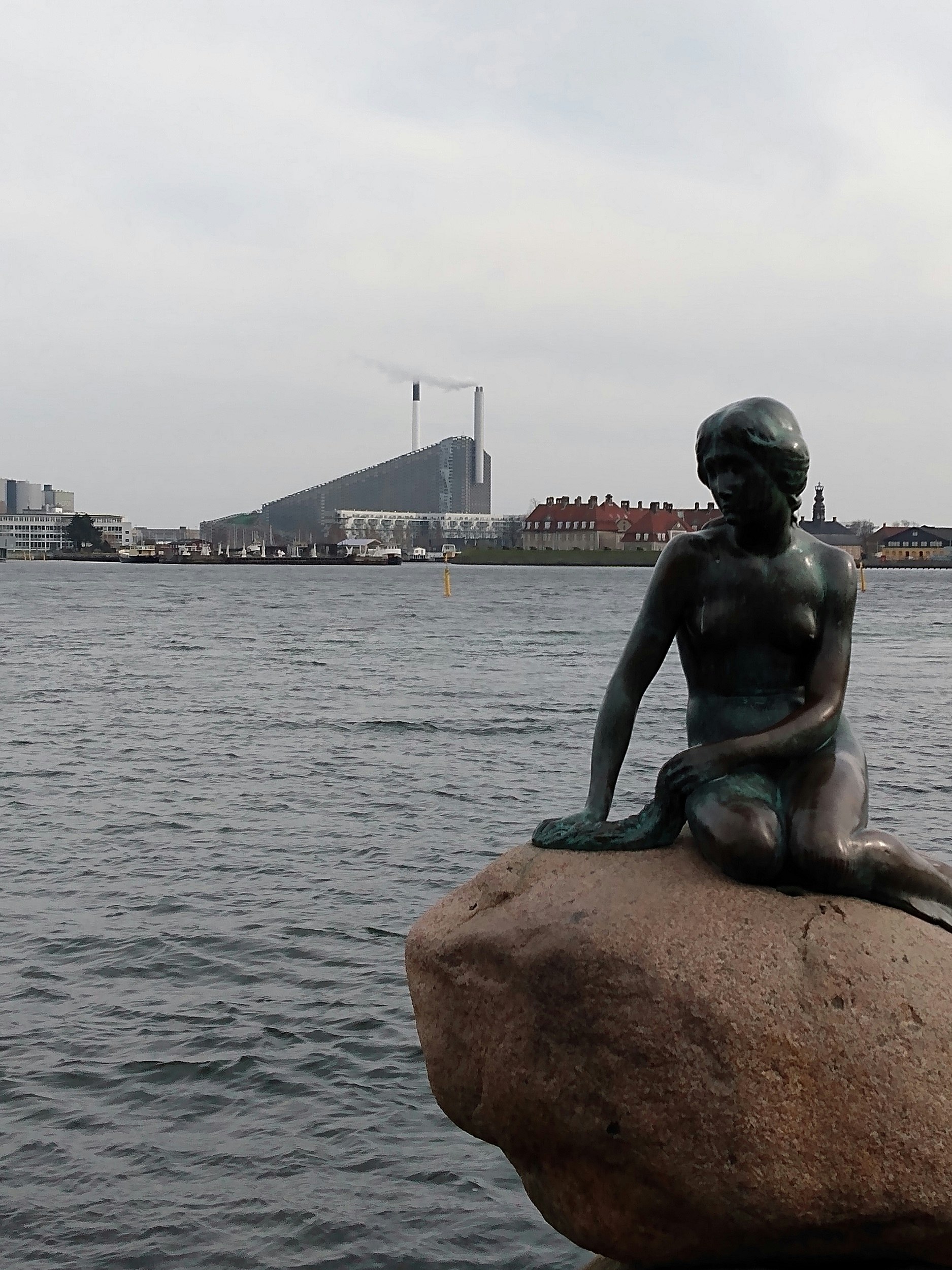 The famous bronze statue of the Little Mermaid sat on a rock by the sea, with a power station with two large chimneys across the water.