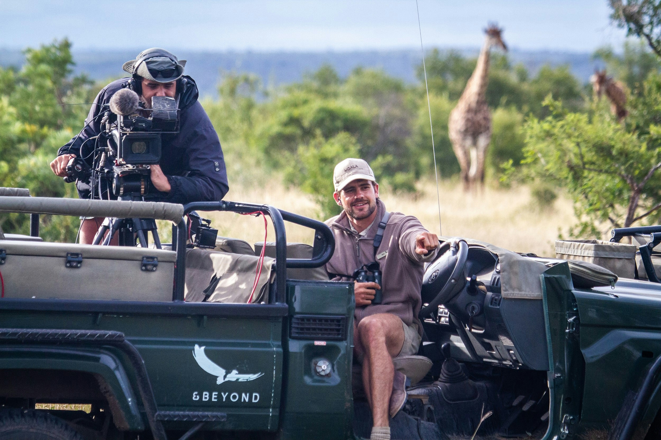 A safari guide sits in an open-topped 4WD and points to the foreground; in the back of the truck is a camera person pointing the lens in the same direction. In the background is a blurred giraffe.
