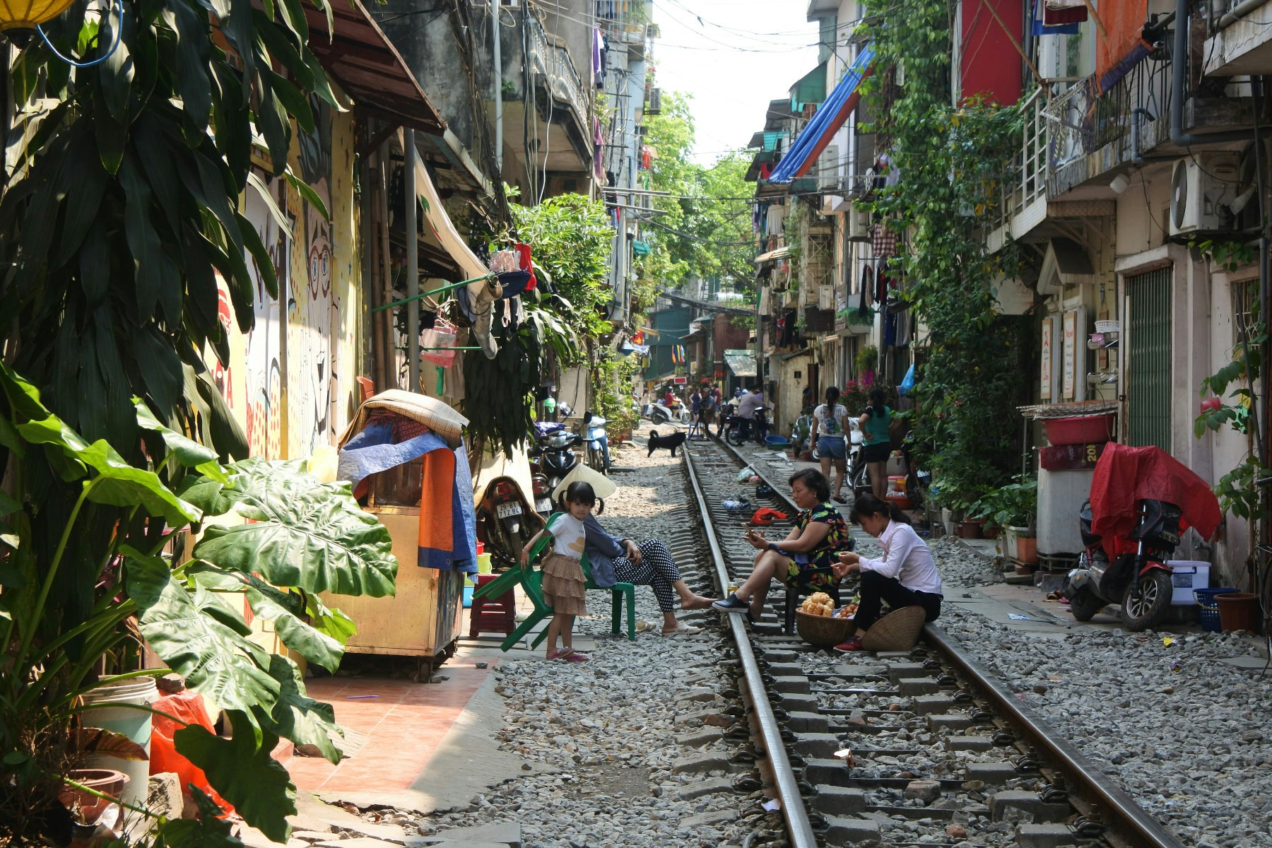 Two women prepare vegetables in the middle of the train tracks while another woman sits and watches a child looks on