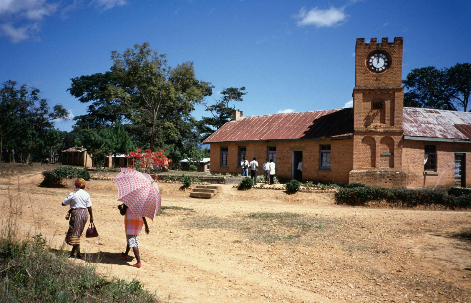 A stone church, with a red tin roof and large castellated clock tower sit beneath blue skies and next to some stands of trees; people mill outside the church, and two women (one holding an umbrella against the sun) walk by on the dirt road in the foreground