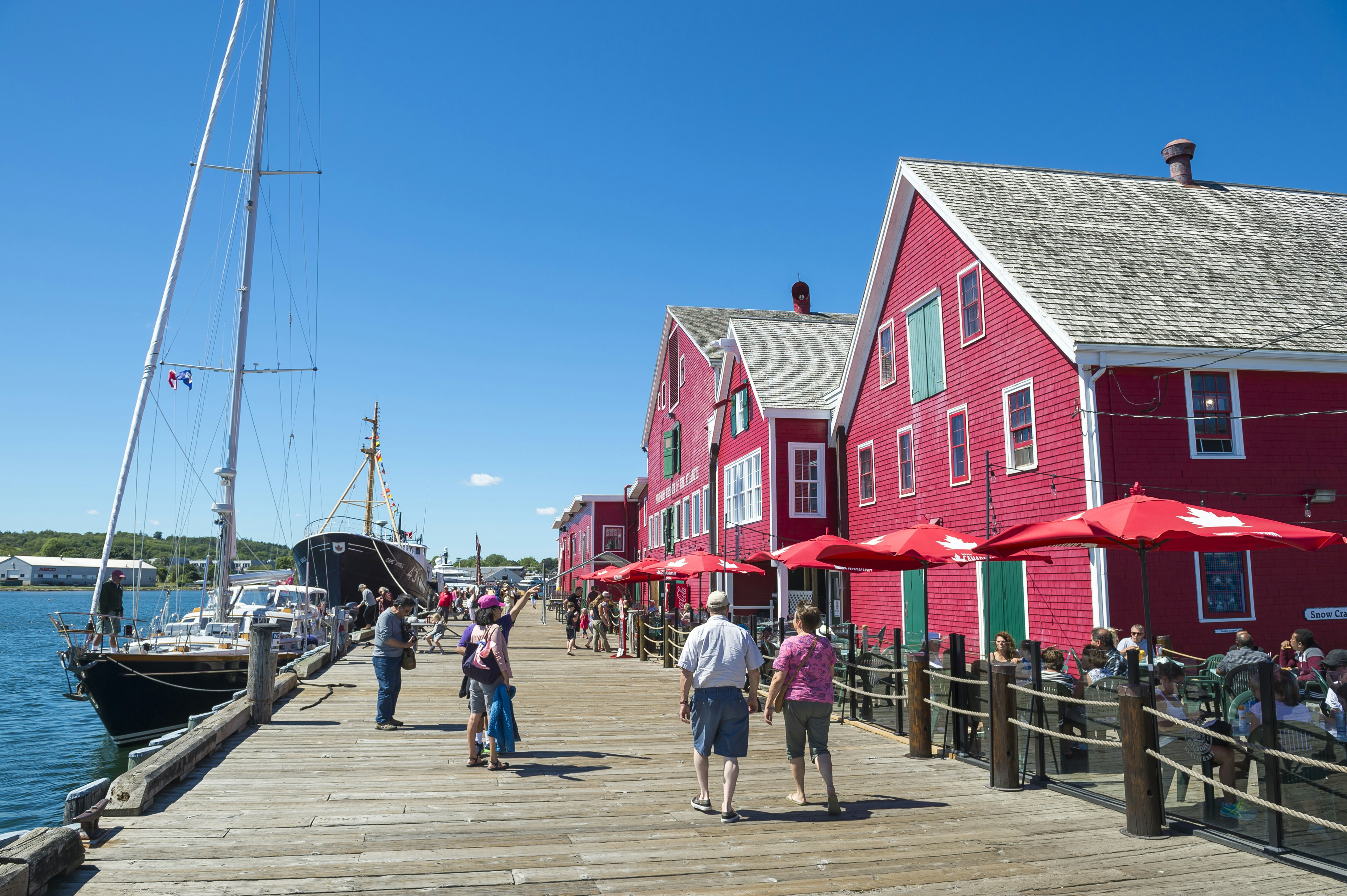 Many people walk down a wooden harbourside promenade; two large boats are moored in the water, and the red, clapperboard buildings of the Fisheries Museum of the Atlantic face the water.