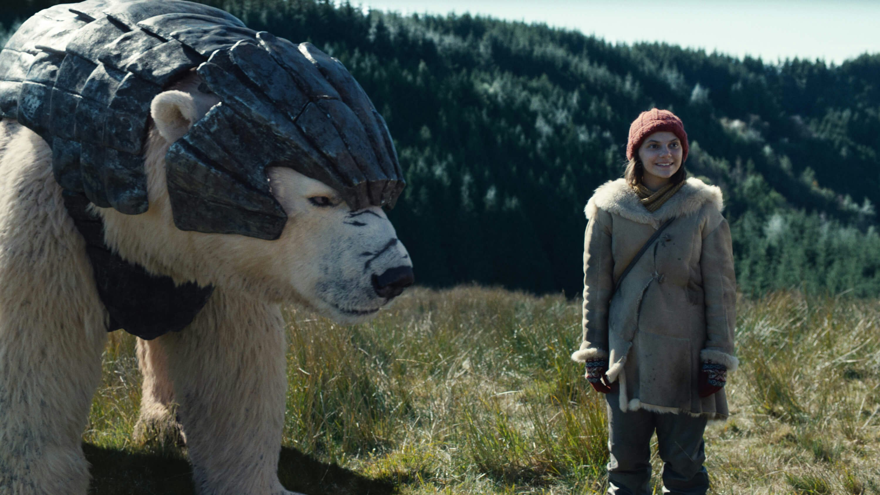 A polar bear with armor stands on the left looking at Lyra, a young girl wearing a red hat, gloves and thick coat.