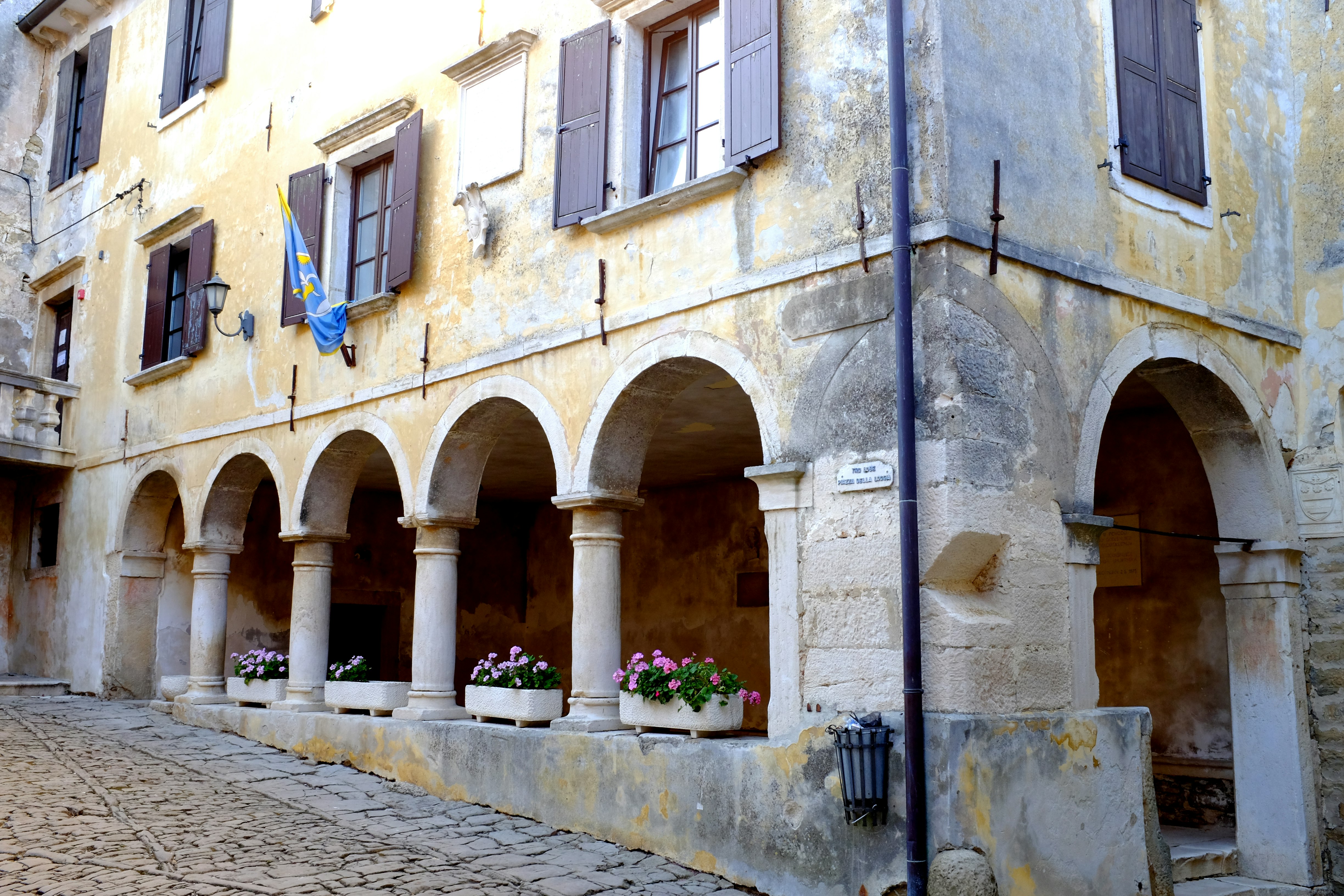 The weathered grey and yellow exterior of one of the loggias in Groznjan on the Croatian border feature window boxes with pink flowers in the archways just above street level, brown shuttered windows, and a blue and yellow flag twisted around the pole