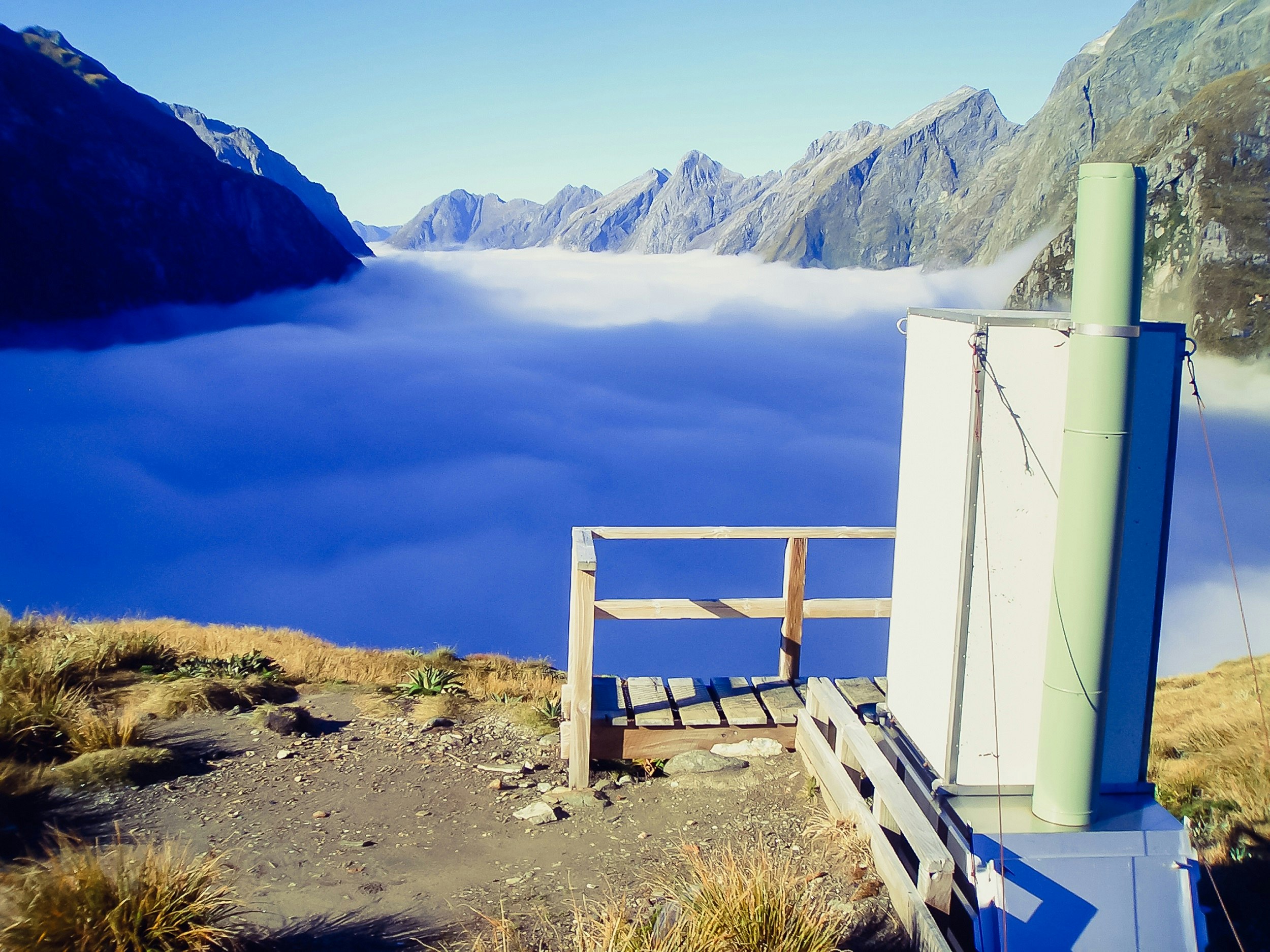 A toilet on top of MacKinnon Pass, Milford Track, New Zealand; it overlooks a fog-filled valley lined by jagged peaks.