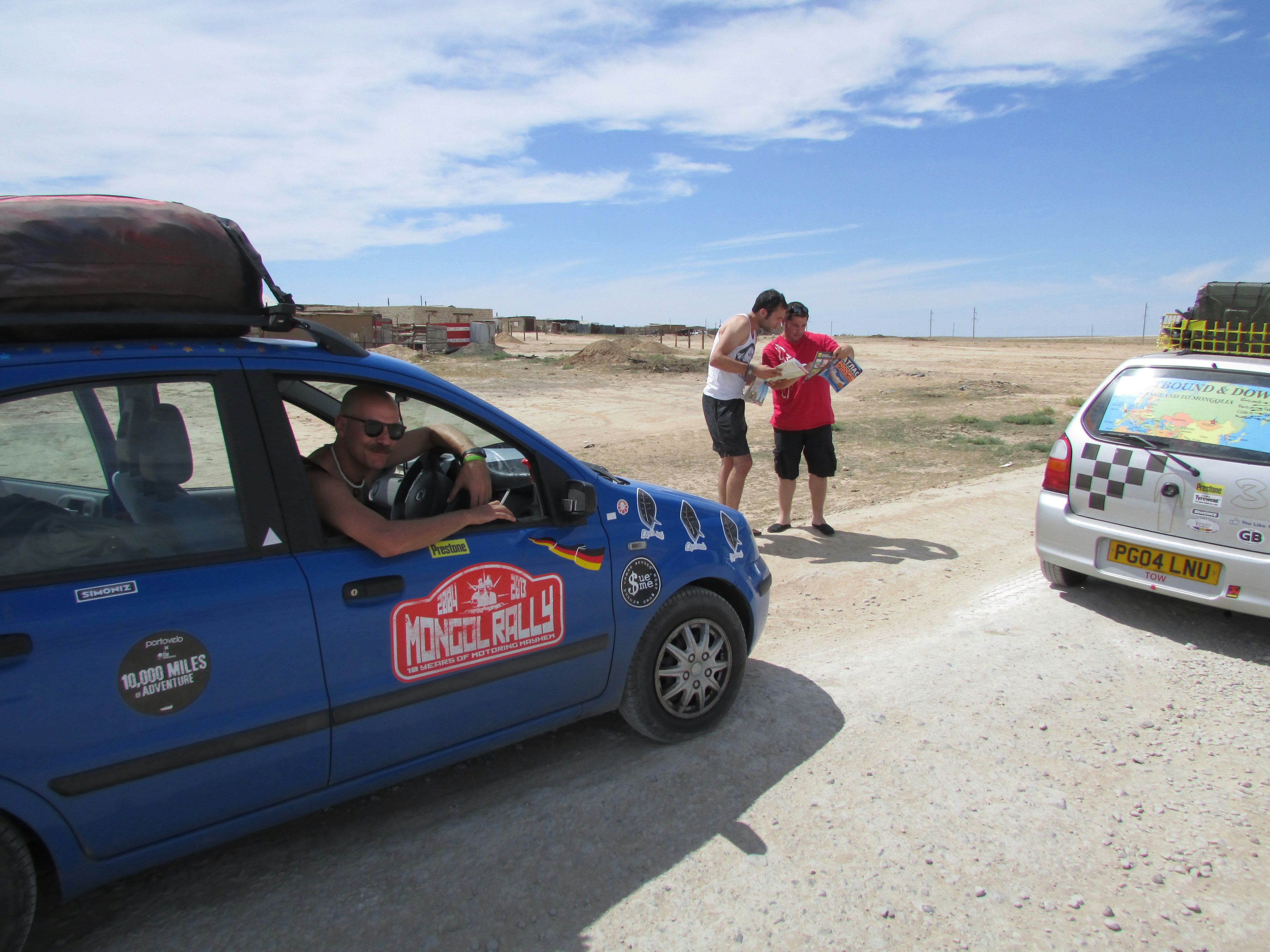 A blue European car is parked next to a white hatchback with a rear window wrap with a map printed on it. Between the cars stand two Mongol Rally participants studying a map. The landscape all around is a bare desert.