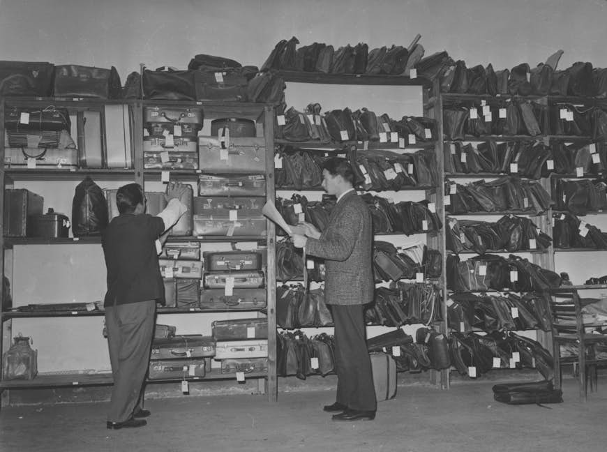 Ett par flygplatsanställda kontrollerar pappersarbete och etiketter relaterade till outlöst bagage på ett kontor för hittegods i Rom 1964.