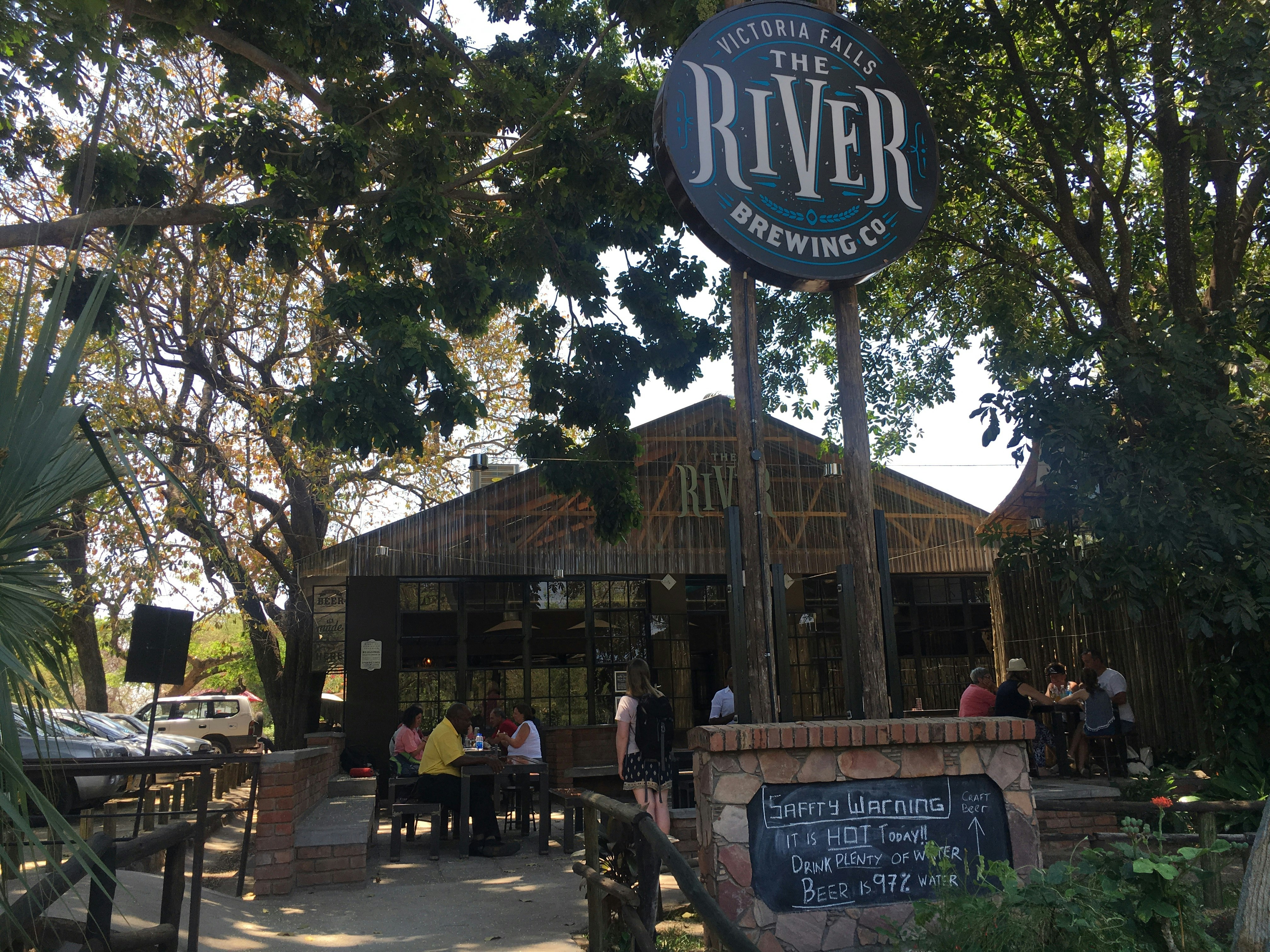 Beneath a tree canopy sits a wooden A-frame building that is the home of The River Brewing Co; people sit around tables sitting in front of the building.