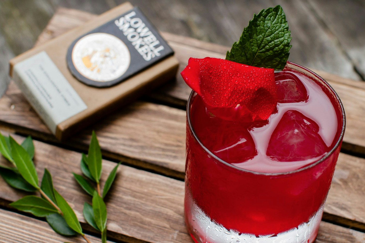 A red-coloured cocktail, decorated with a rose petal, and a pack of Lowell Smokes in the background