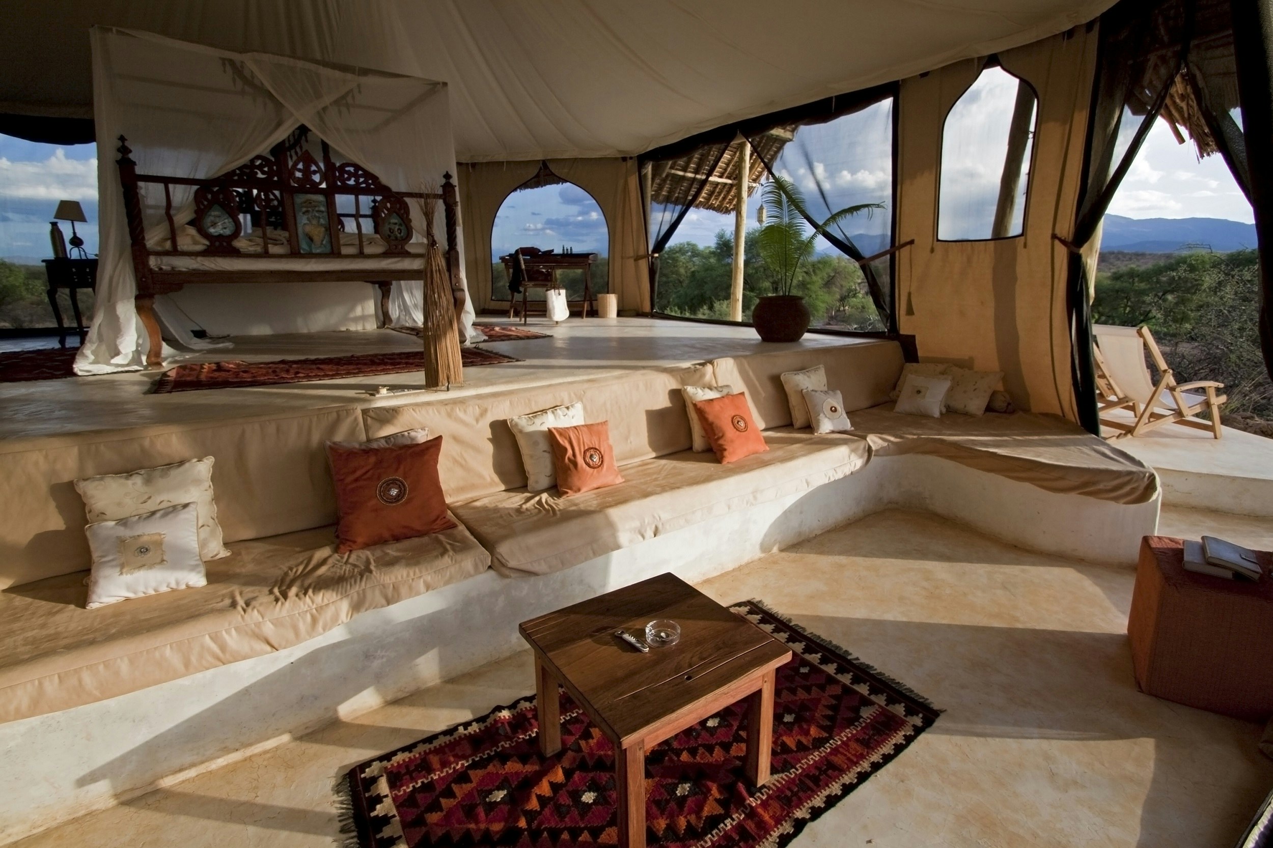 The vast, multi-levelled interior of a safari tent in Kenya; polished tan-coloured concrete floors with rugs are surrounded by a built-in seating area, which flows onto an upper level with a four-poster bed.