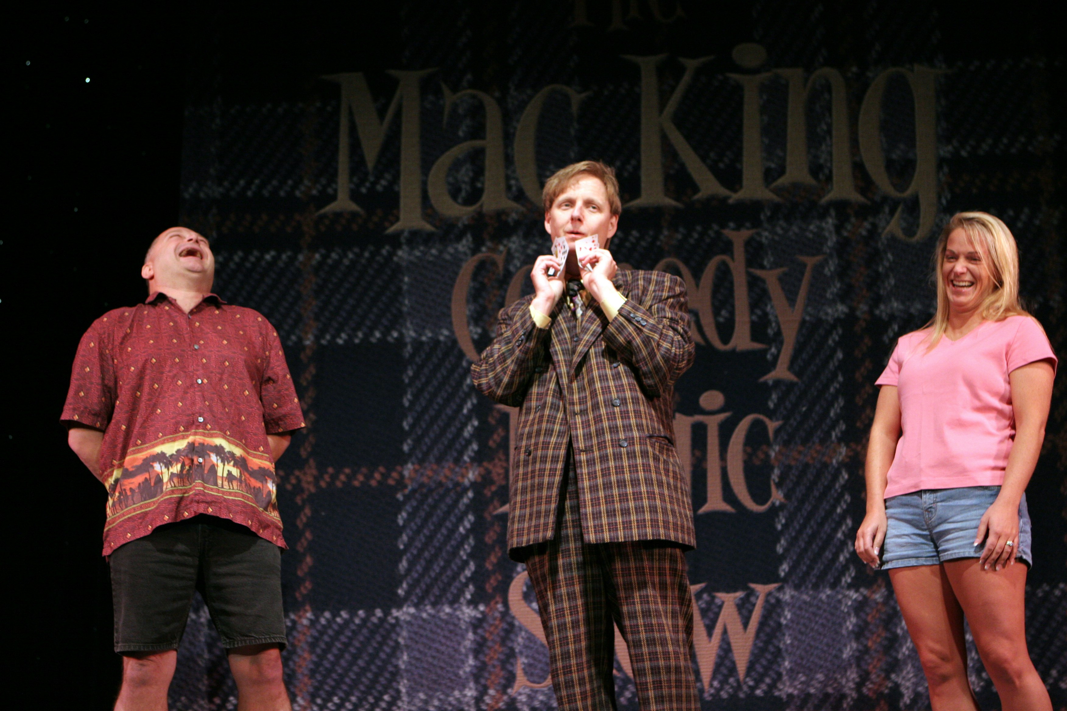 Magician and comedian Mac King performs a card trick on stage at Harrah's in Las Vegas, while a man and a woman stand either side of him, laughing.
