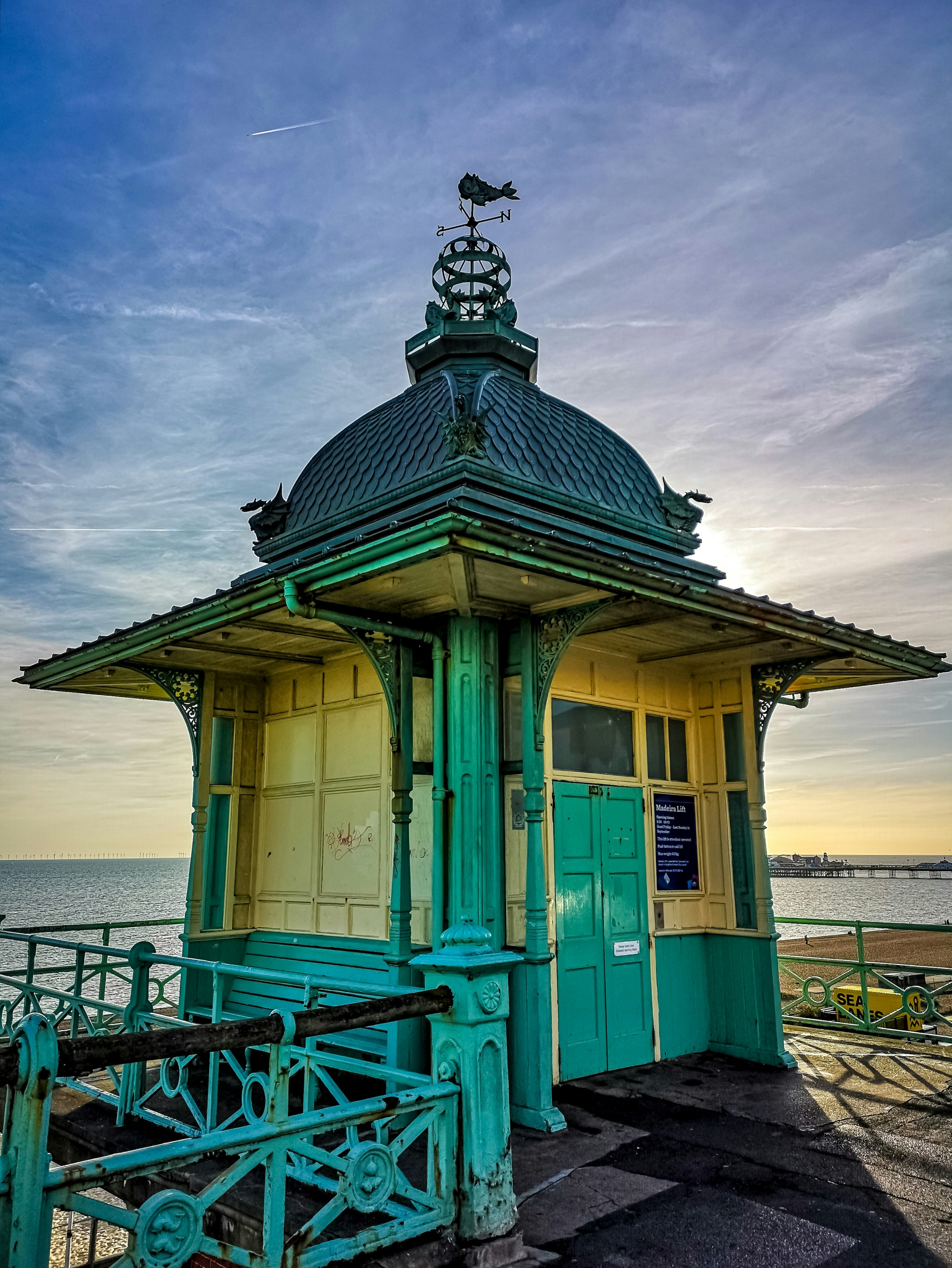 A Victorian elevator standing on the Brighton seafront. It is painted buttermilk yellow and teal blue, and it's topped with a dome and a weather vane.