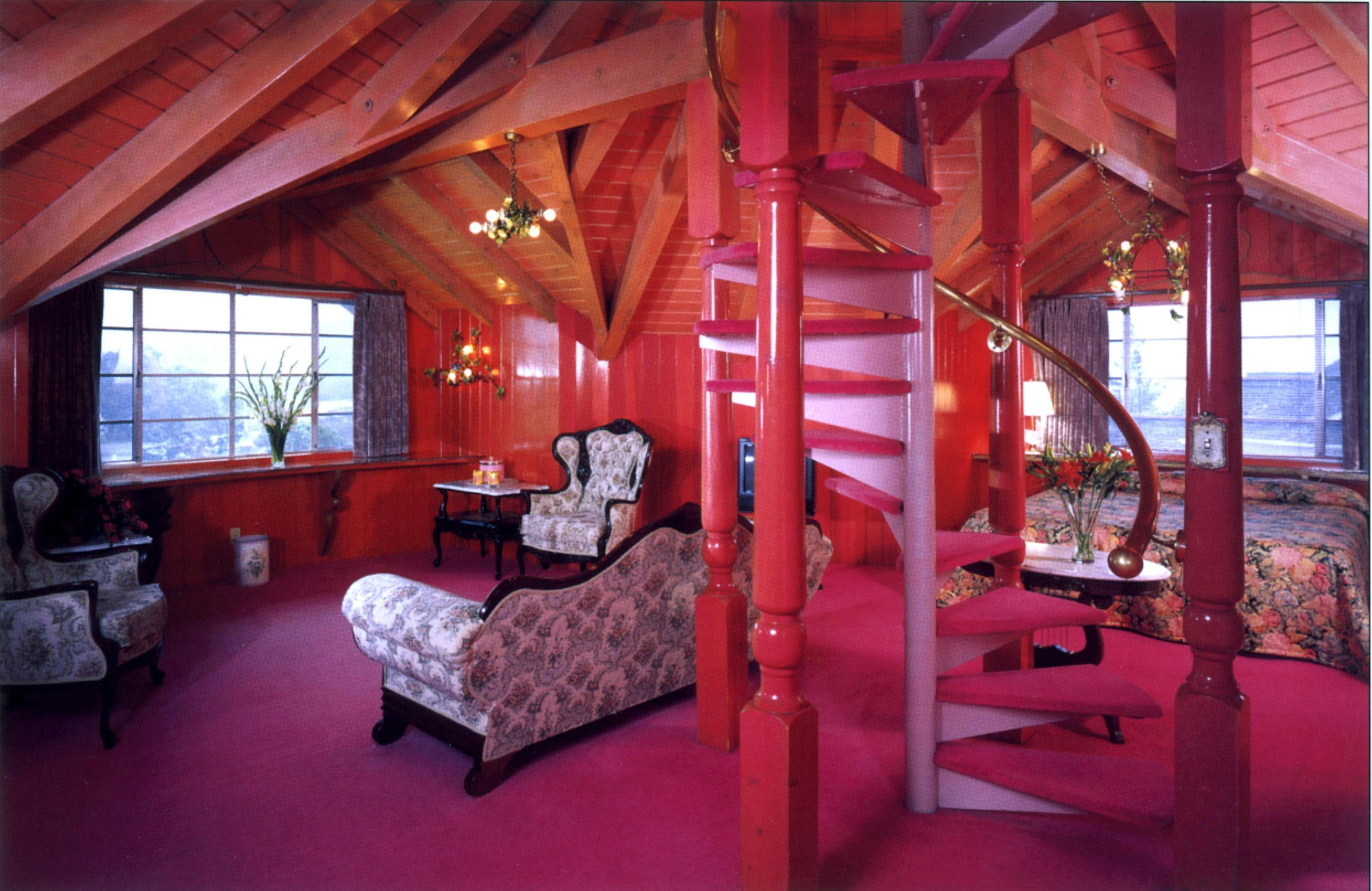 The bright pink interior of the Love Nest room at Madonna Inn has angled ceilings from the whimsical hotel's exterior turrets, and a large central pink spiral staircase. The bed is by a window to the right, while to hte left is a seating area with white floral China patterned camel back Victorian sofa and a matching wingback chair 