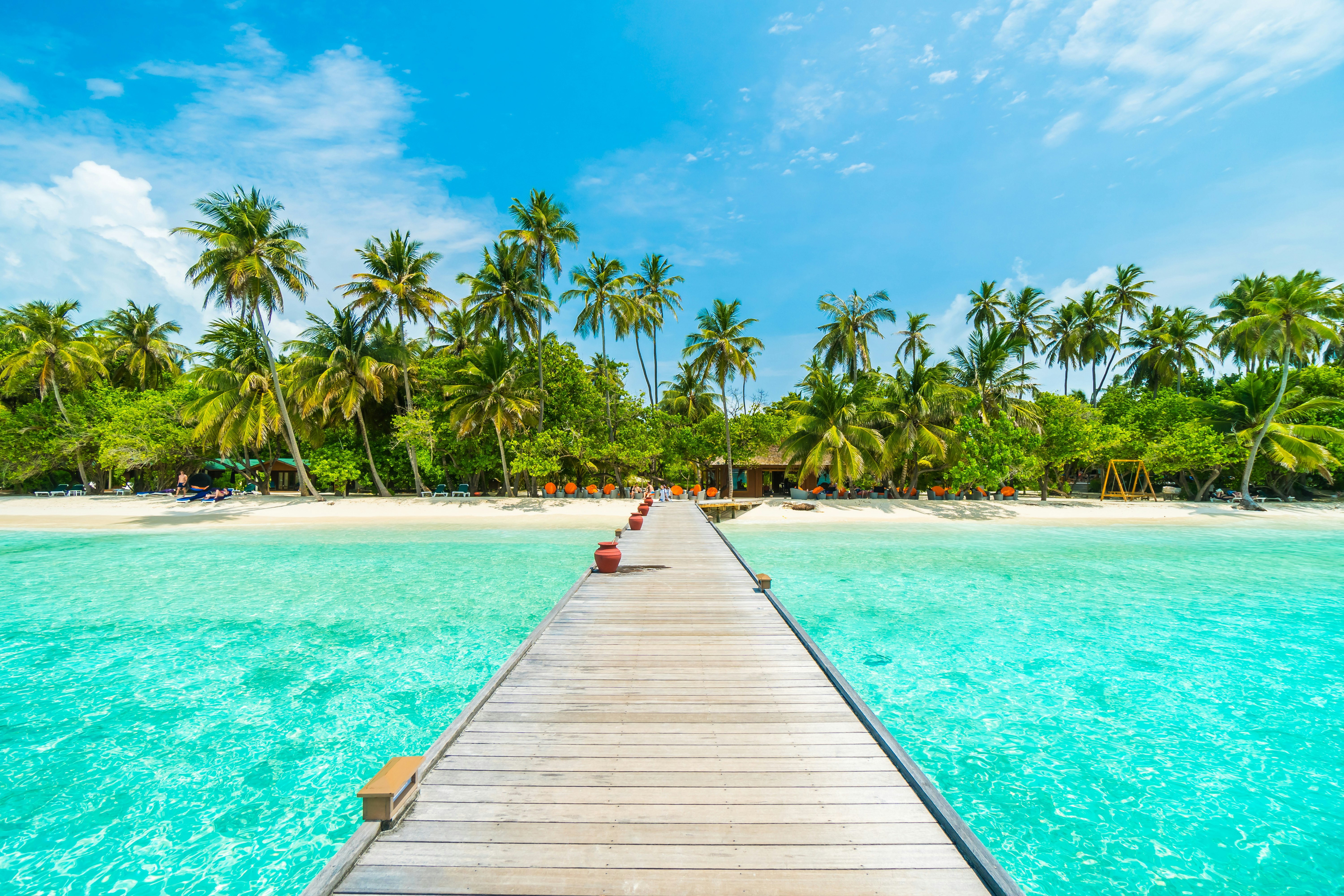 Maldives island resort with jetty, beach, sea and coconut palm trees on blue sky.