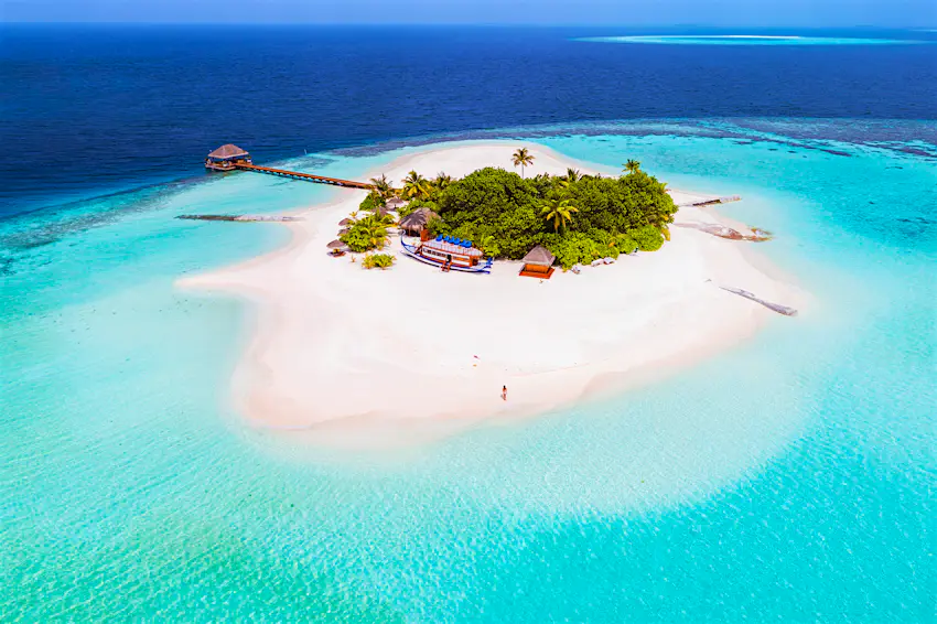 Maldives Ari Atoll An aerial shot of a small sandy island covered with palm trees. There's a wooden pier stretching out to the reef on the edge of the island
