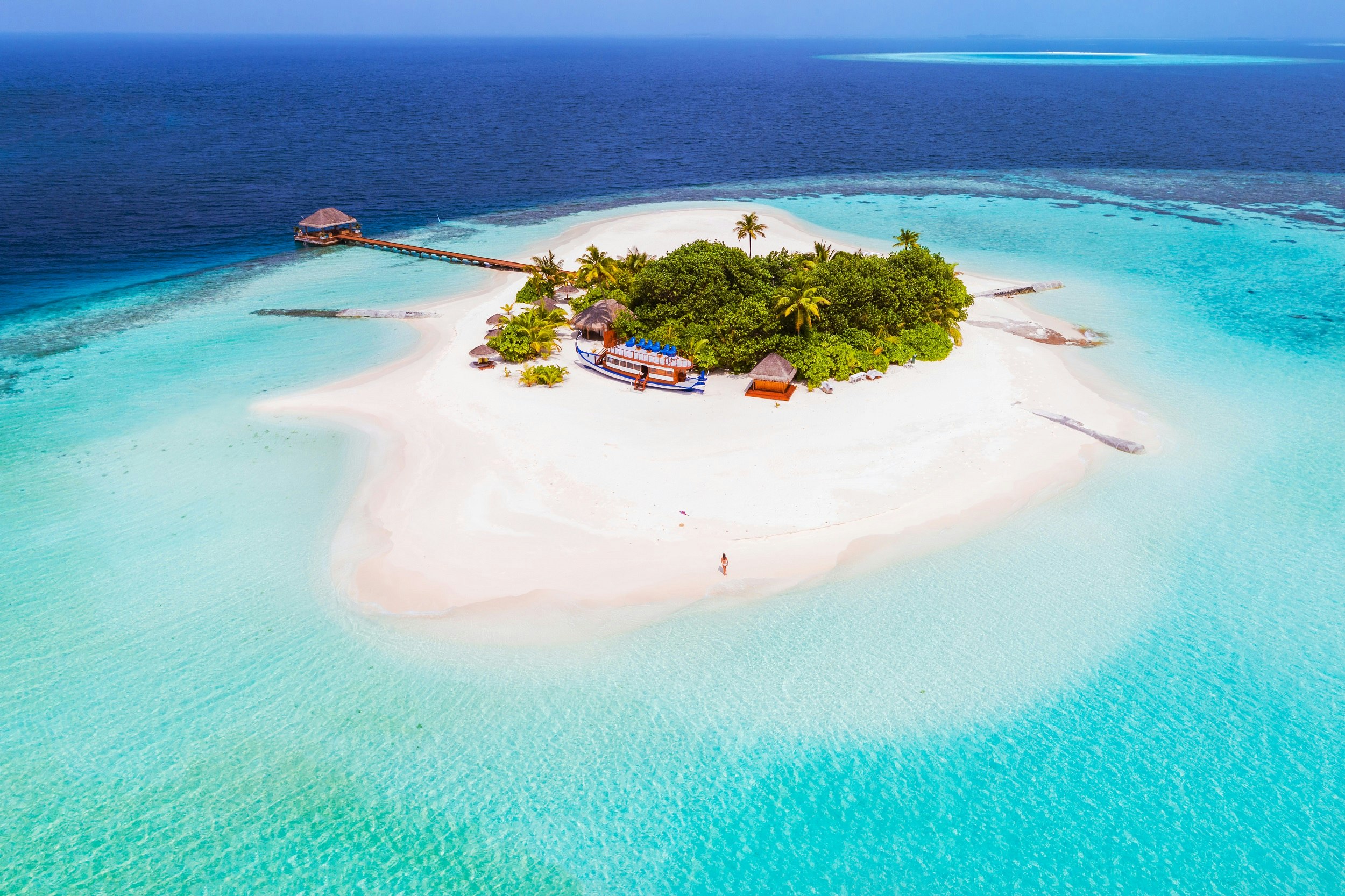 An aerial shot of a small sandy island covered with palm trees. There's a wooden pier stretching out to the reef on the edge of the island