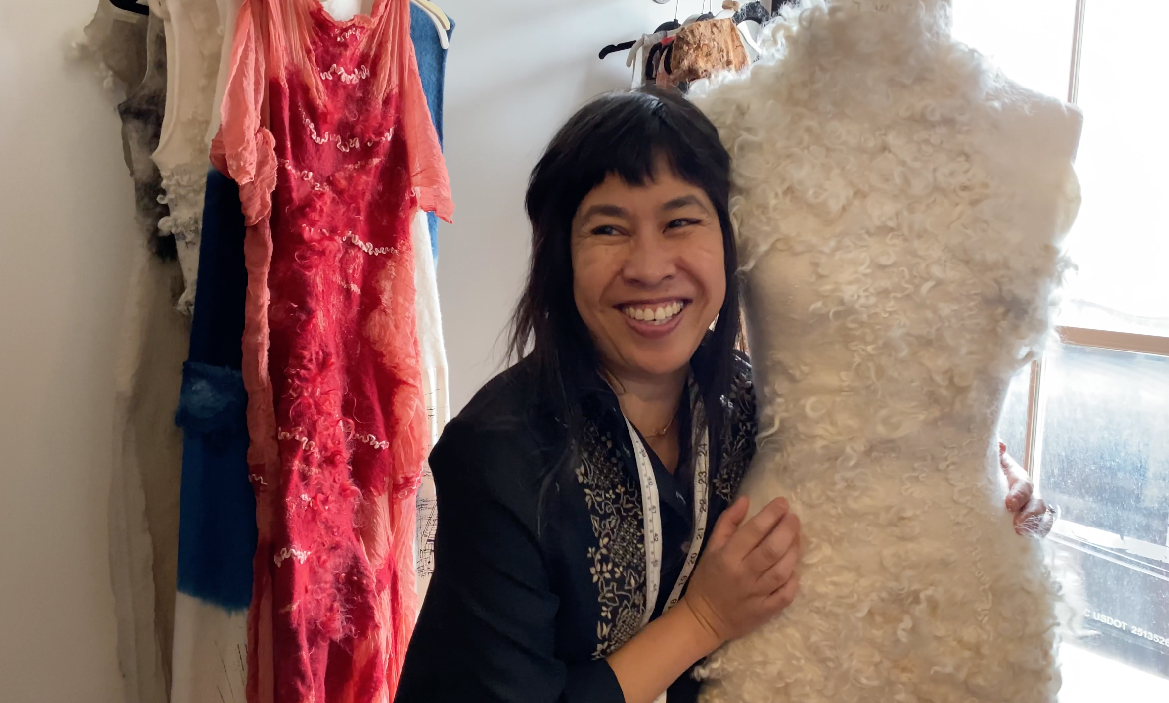 Celeste Malvar-Stewart poses with one of her custom wedding gowns, hugging it and the dress form it's on. She has short black hair and a big smile on her face, with a white tape measure draped around her neck. Behind her are several more of her dresses, including a bright red dress with a swirl of curly wool down the front